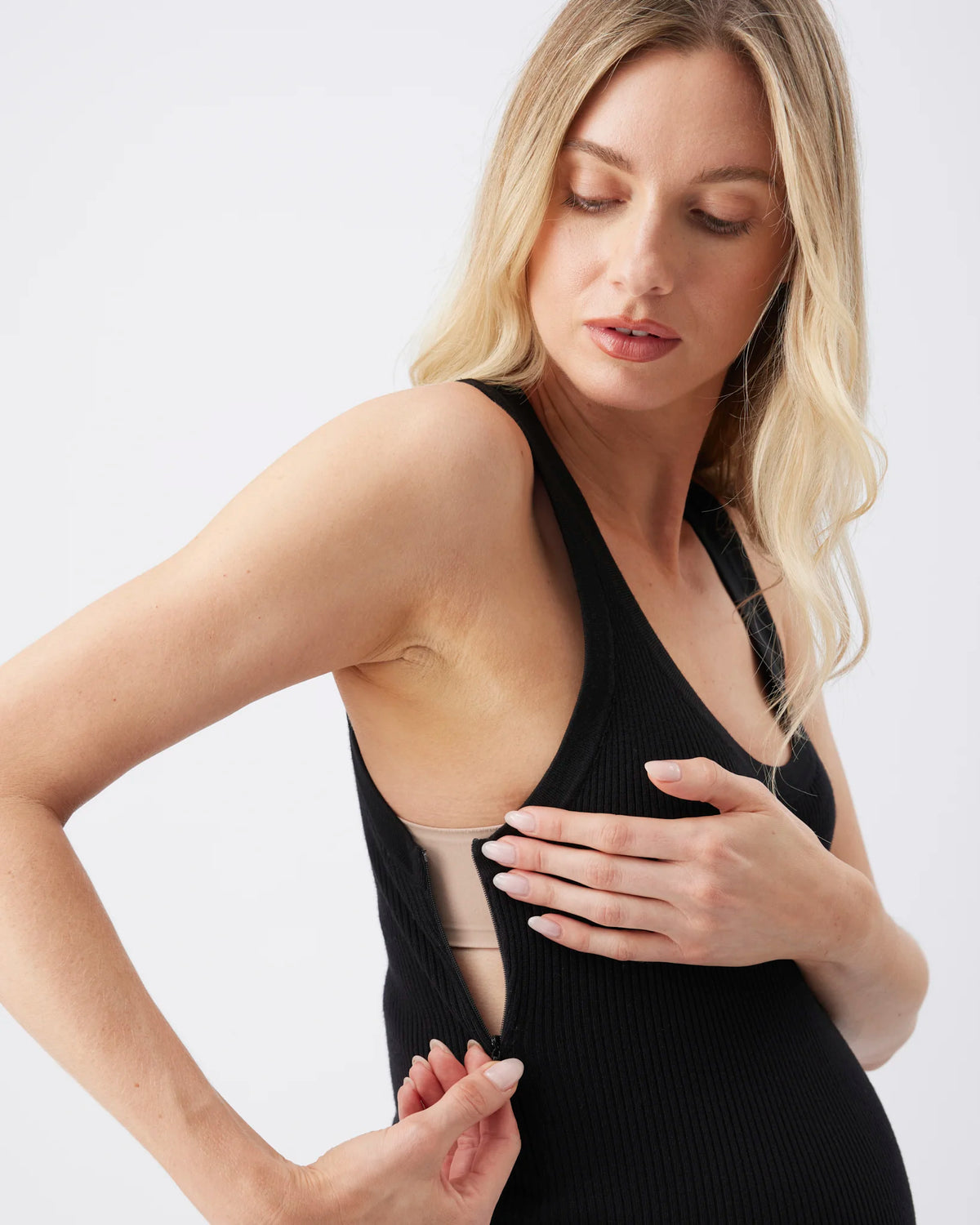 A blonde woman wearing a black ribbed maternity tank top with a side cutout, holding a beige nursing bra underneath to demonstrate the product's function, against a clean white background.
