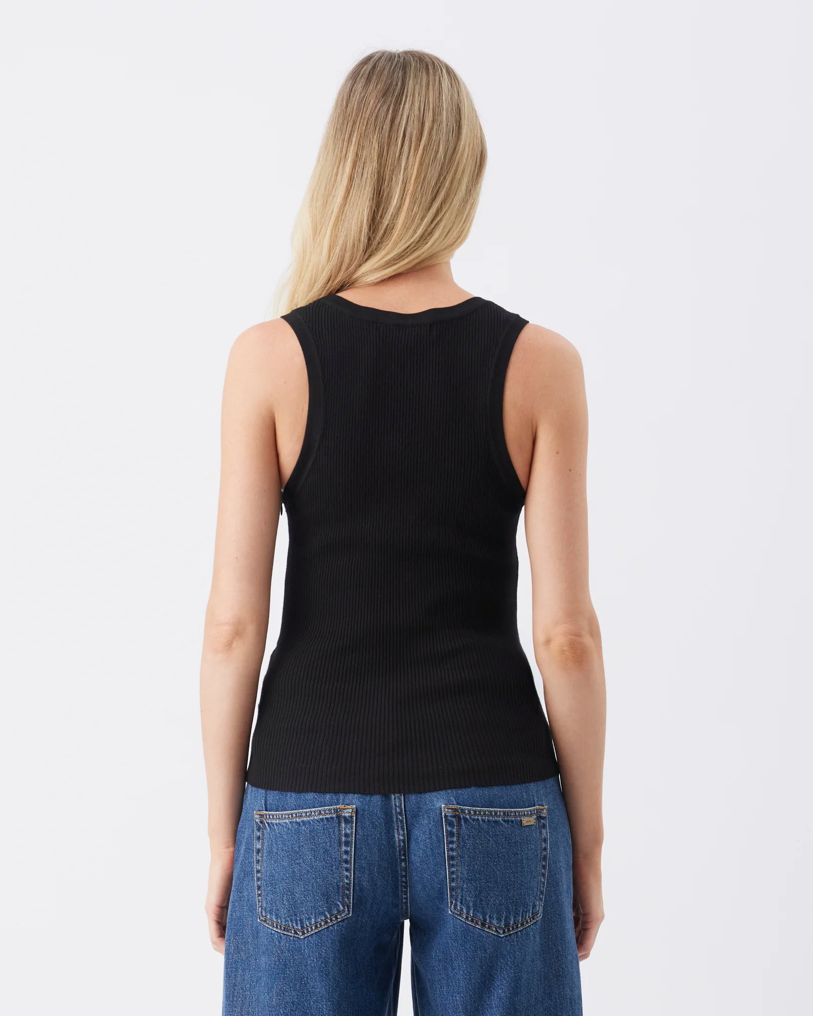 A woman viewed from the back wearing a black sleeveless ribbed knit top and blue jeans against a plain light gray studio background.