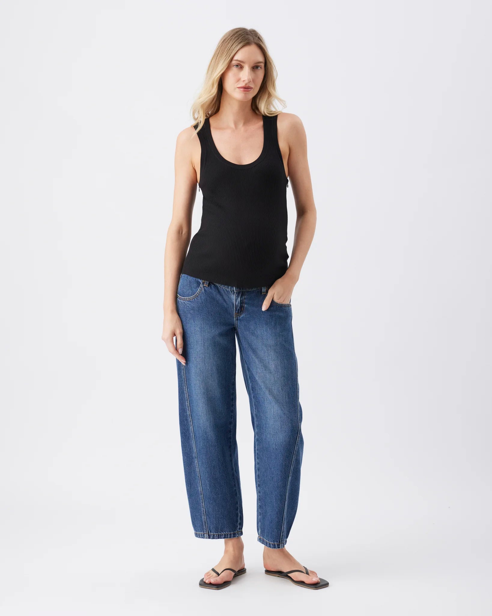 A woman modeling a black sleeveless scoop-neck tank top and wide-leg blue jeans with brown sandals, standing against a plain white studio background.