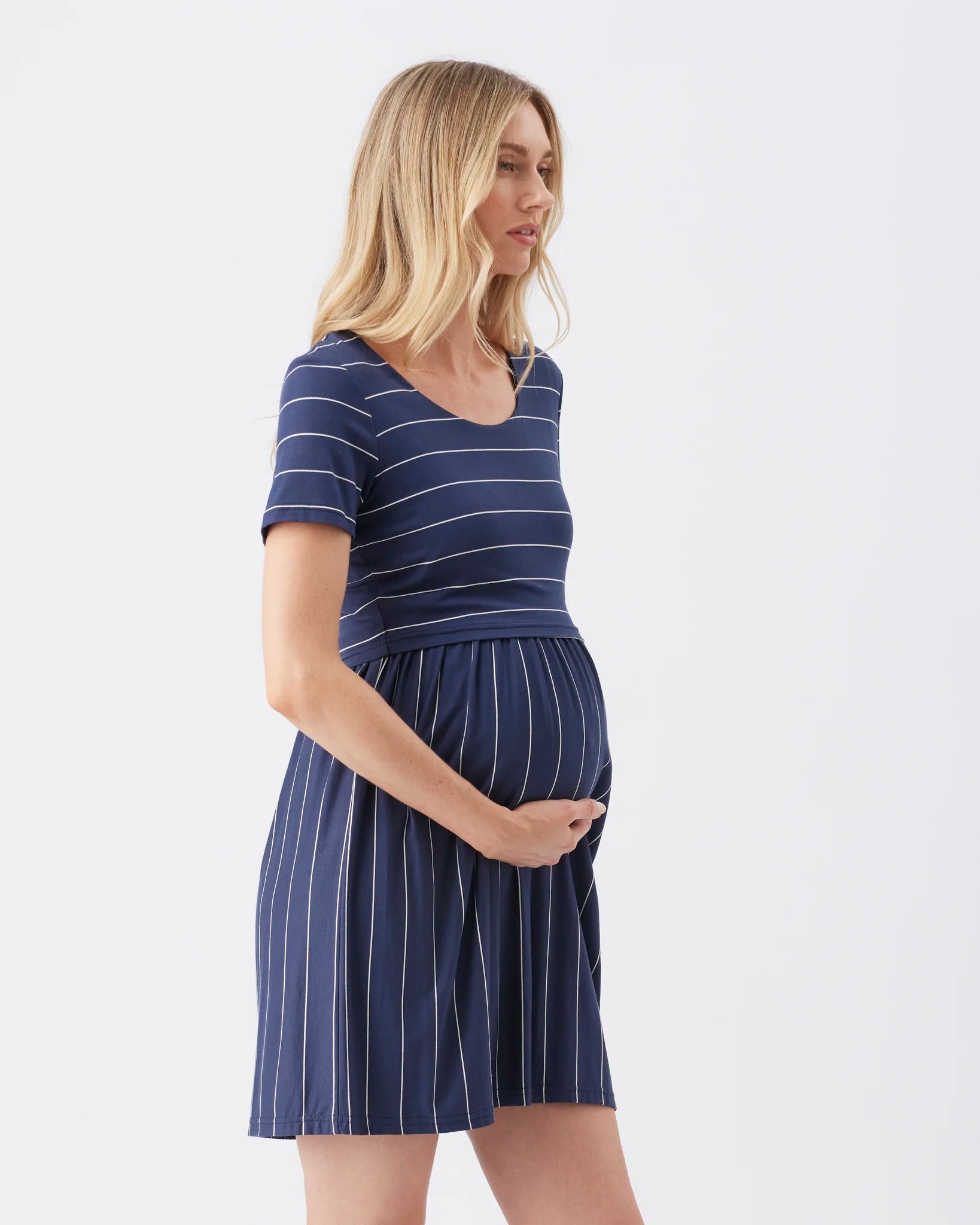 A pregnant woman wearing a short-sleeve maternity dress with navy blue and white vertical stripes, standing against a plain white studio background.