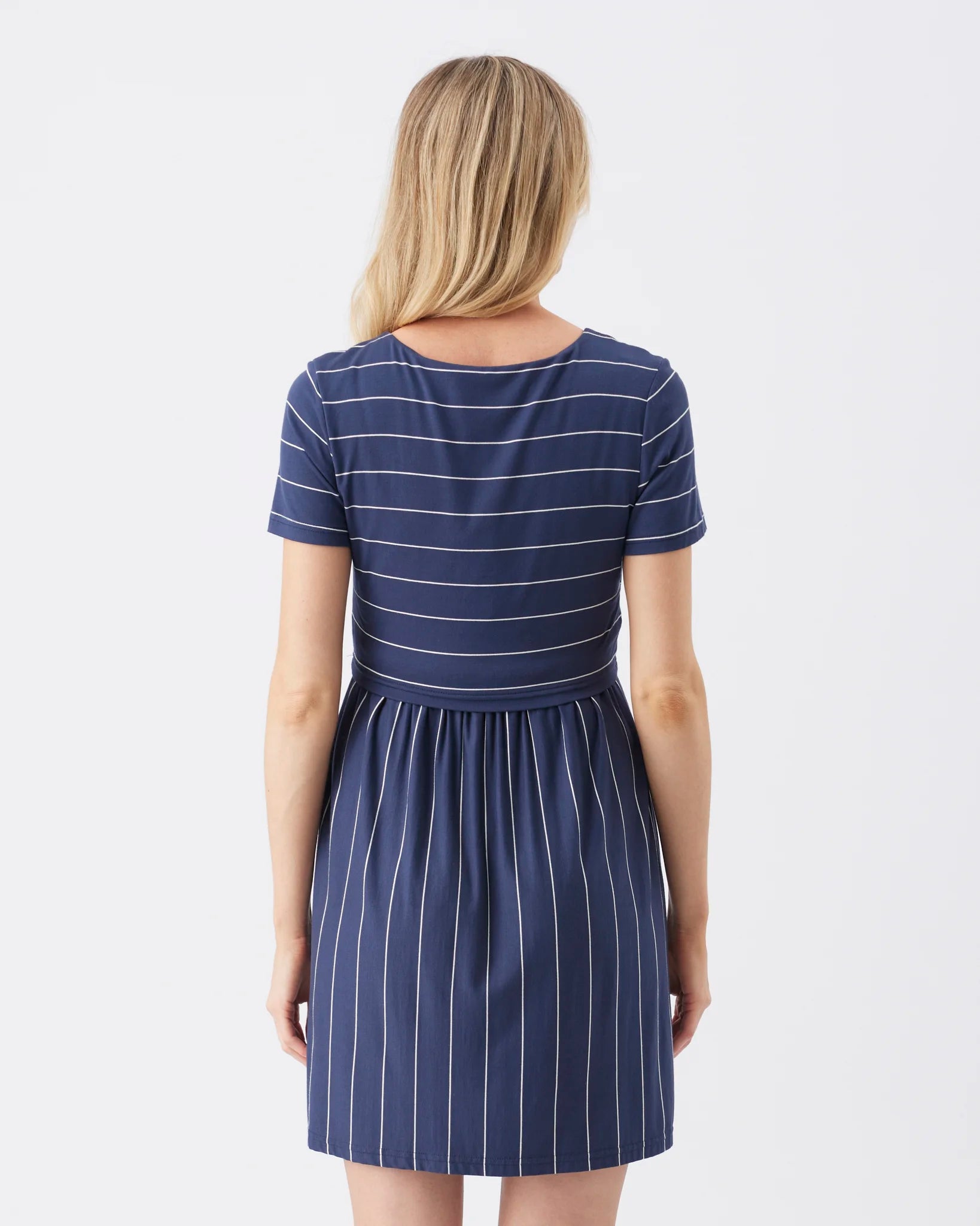 A woman viewed from the back wearing a short-sleeve navy dress with thin white horizontal stripes on the bodice and vertical white pinstripes on the skirt, against a plain light gray studio background.