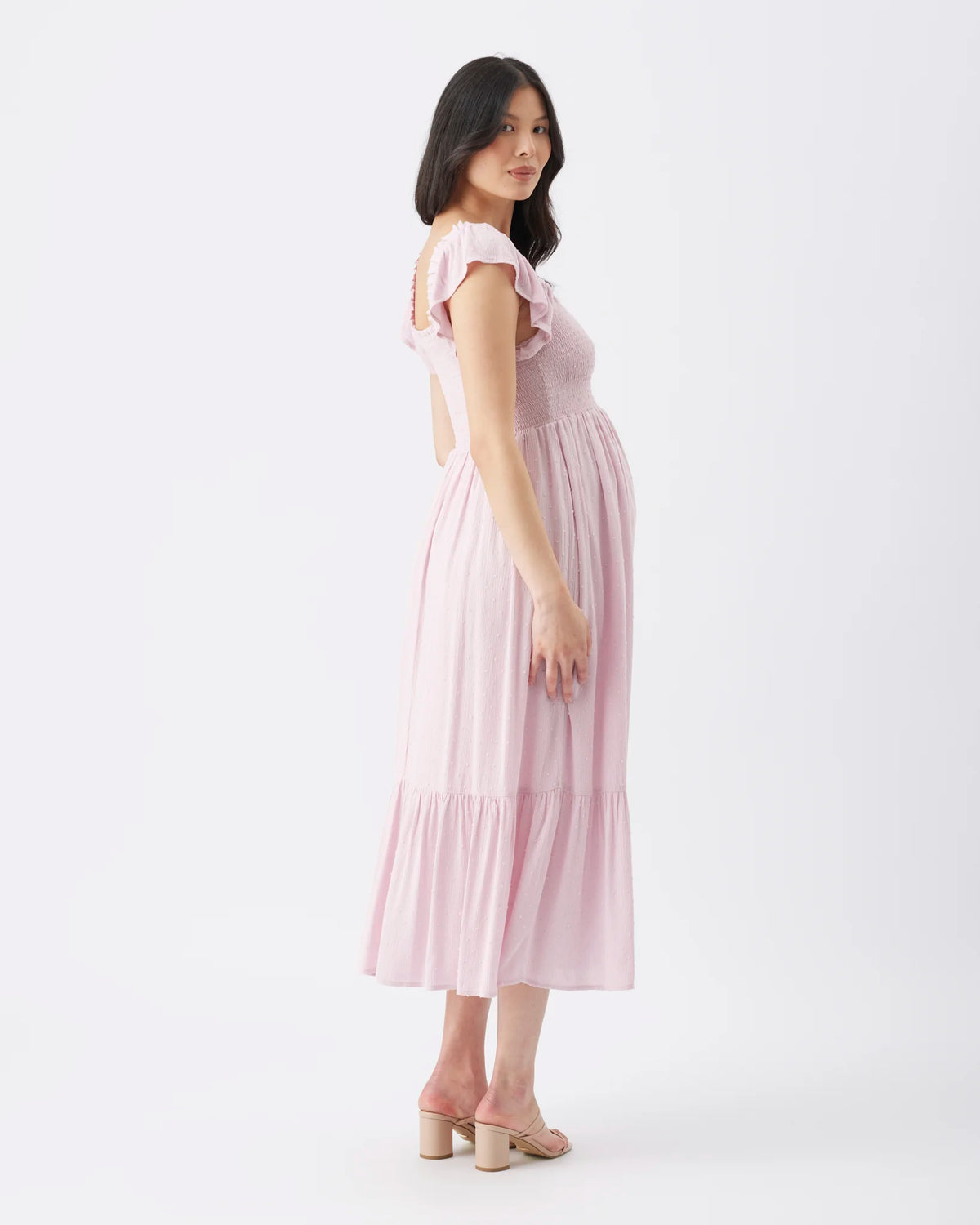 A pregnant woman wearing a light pink tiered midi dress with ruffled shoulder straps and a backless cut, standing against a plain white studio background.