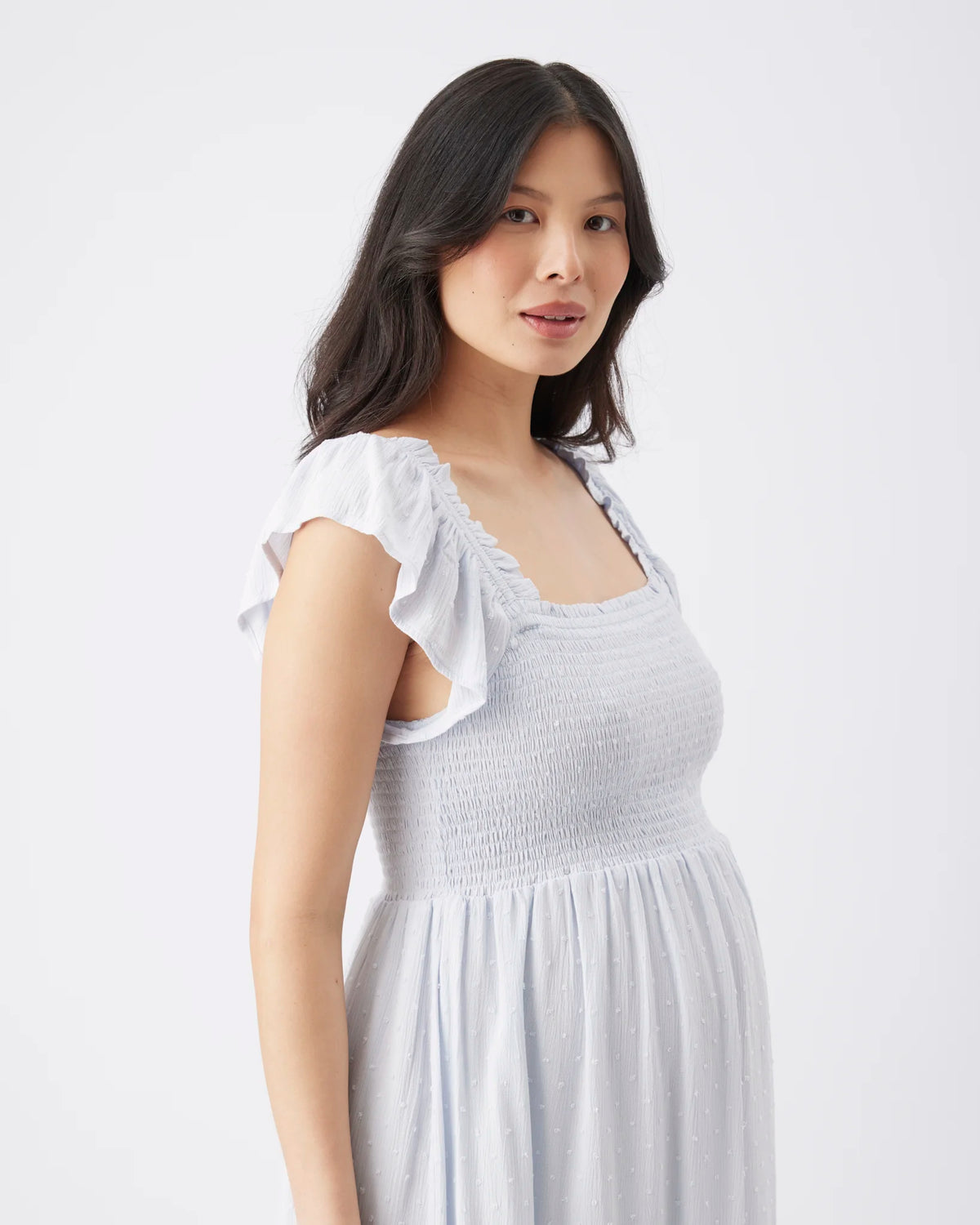 A woman wearing a white smocked dress with ruffled cap sleeves, photographed against a clean light gray studio background.