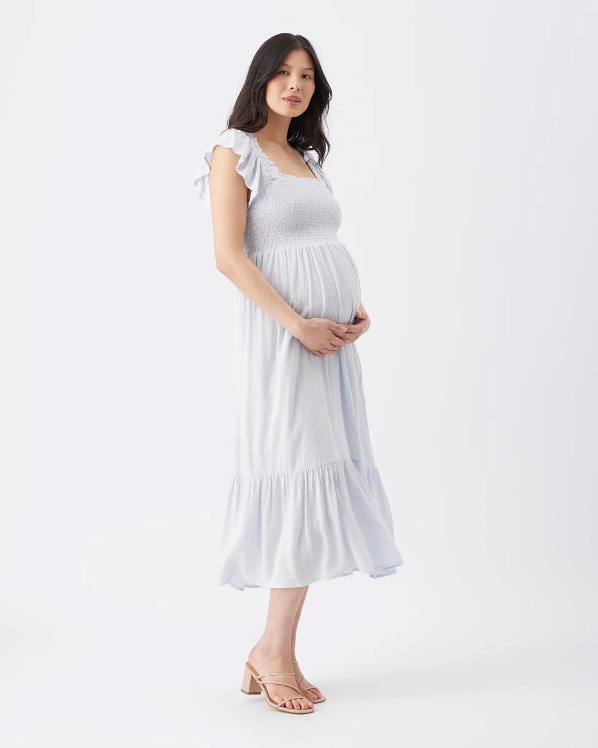 A pregnant woman wearing a light blue sleeveless midi maternity dress with ruffled shoulder straps and a tiered skirt, standing against a plain white studio background.