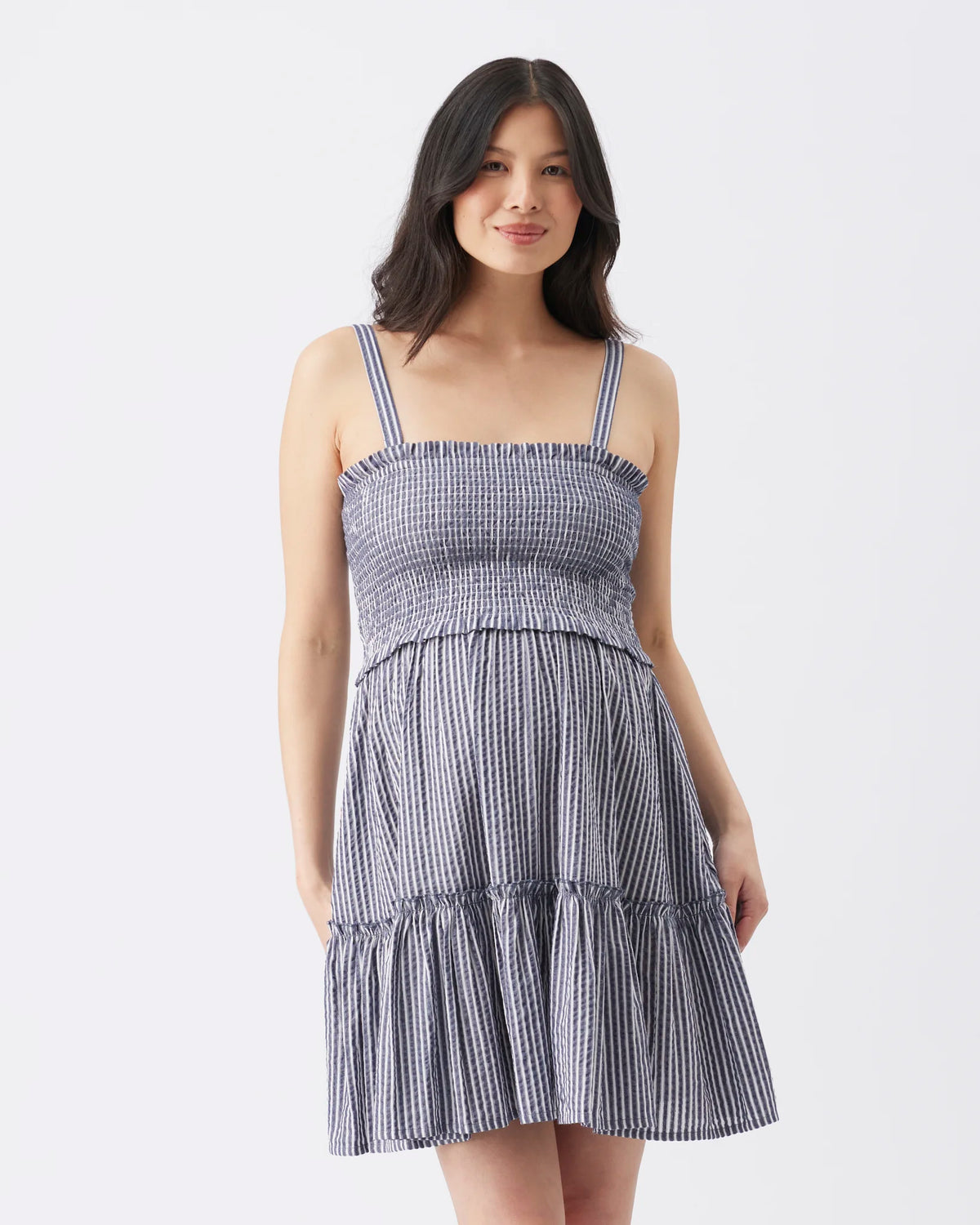 A woman modeling a sleeveless tiered dress with a smocked bodice and thin shoulder straps, featuring a blue and white vertical stripe pattern, against a plain white studio background.