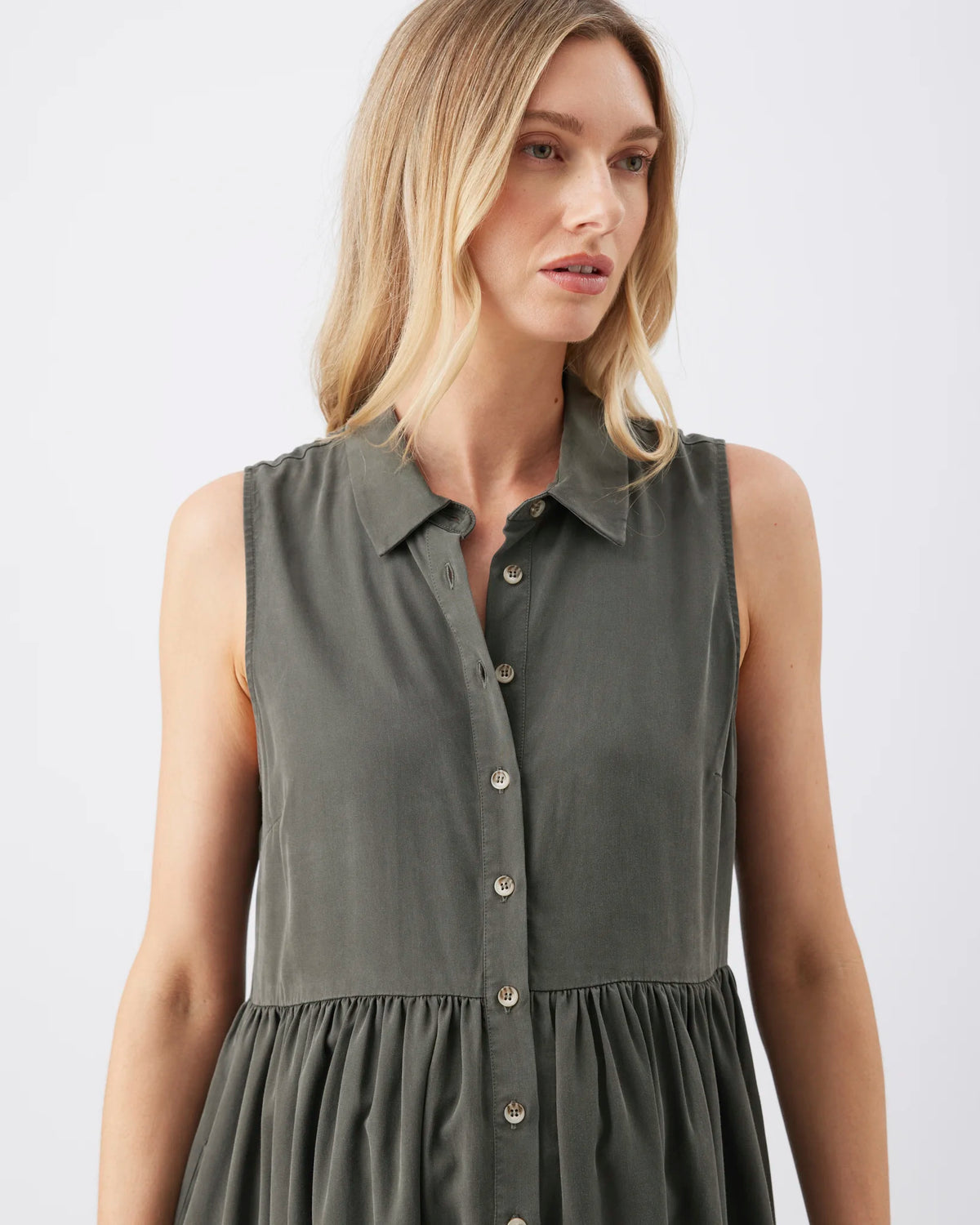 A woman wearing a sleeveless collared button-front dress in a muted olive green color, shown from the shoulders to the hips against a plain light gray studio background.