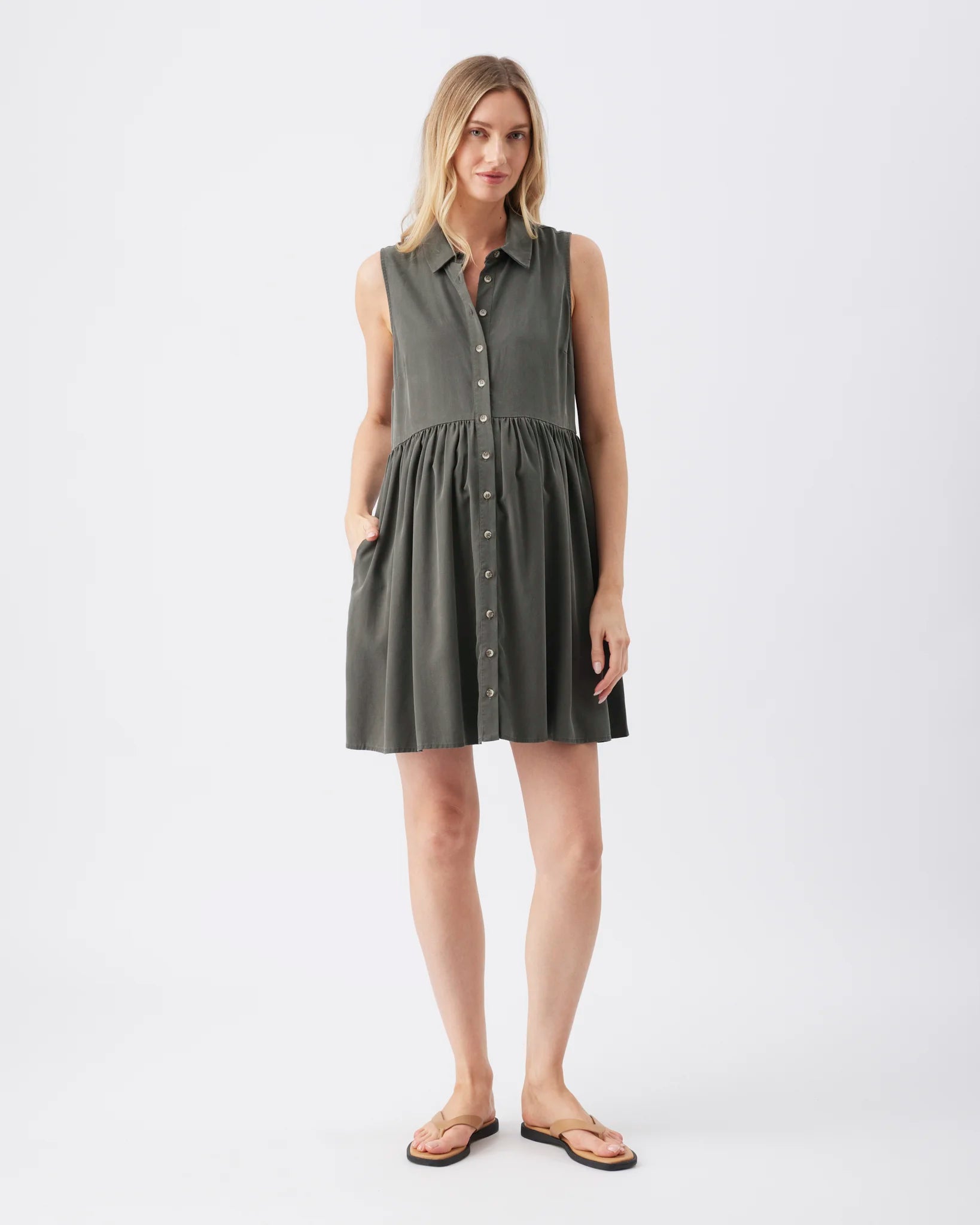 A woman modeling a sleeveless, button-front shirt dress in a dark olive green color, paired with tan flip-flop sandals, against a plain white studio background.