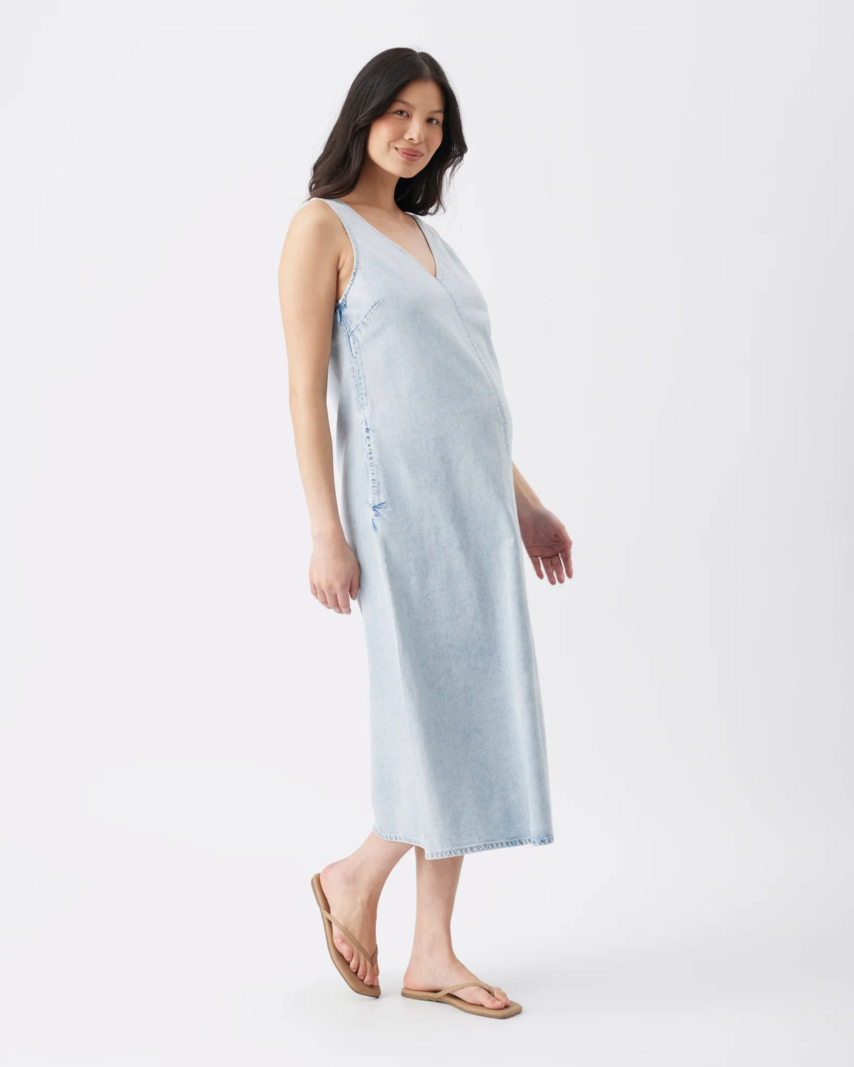 A pregnant model wearing a light blue denim maternity dress and brown flip-flop sandals, standing against a plain white studio background.
