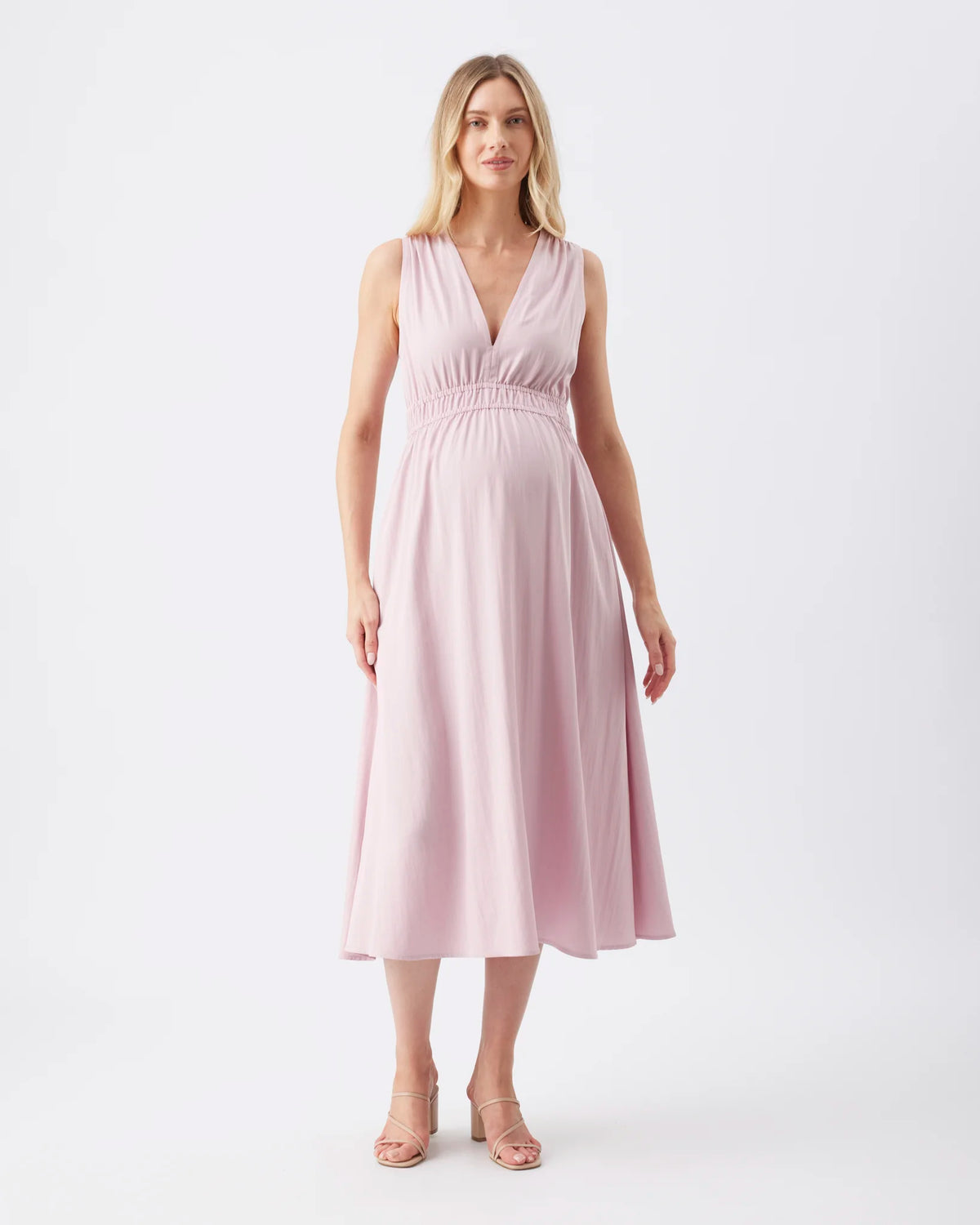 A woman modeling a sleeveless, V-neck midi dress in a soft dusty pink color, standing against a plain white studio background.