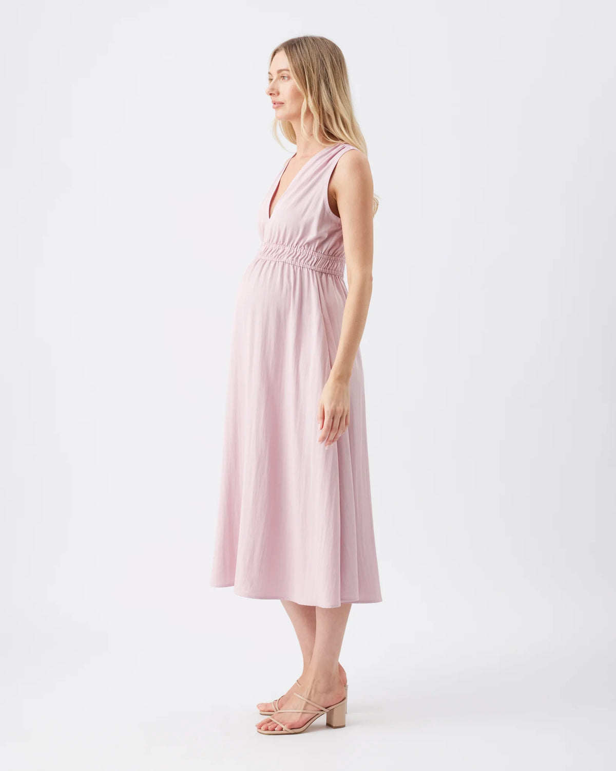 A woman modeling a sleeveless dusty pink midi dress with a smocked waist and V-neckline, shown in profile against a plain white studio background.