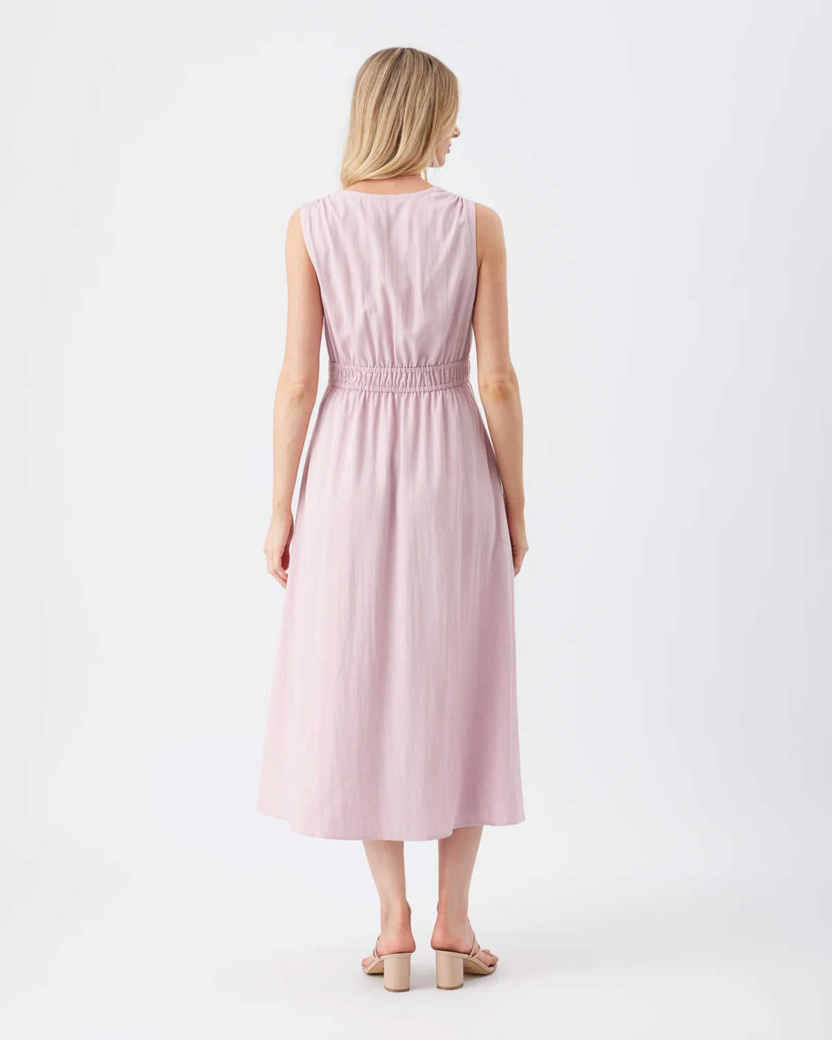 A woman viewed from the back wearing a sleeveless midi dress in a very light blush pink color, paired with beige block-heel sandals, against a plain white studio background.