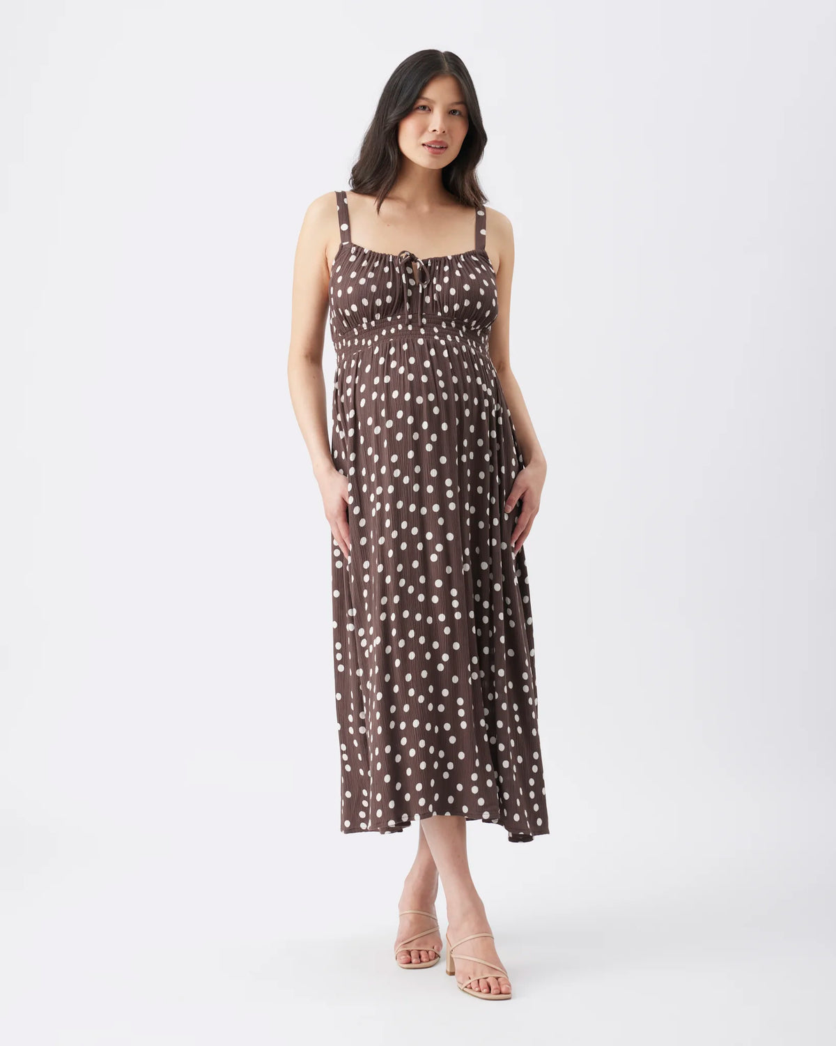 A woman modeling a sleeveless midi dress with a brown base and white polka dots, standing against a plain white studio background.