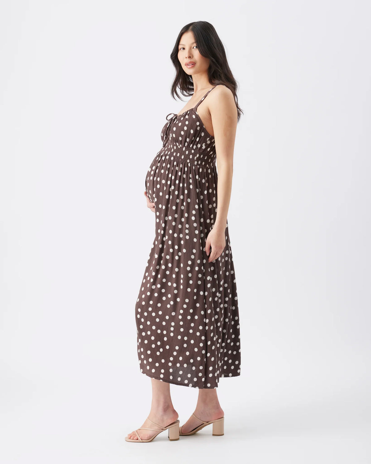 A pregnant woman modeling a brown spaghetti-strap maxi dress with a white polka-dot pattern, standing against a plain white studio background.
