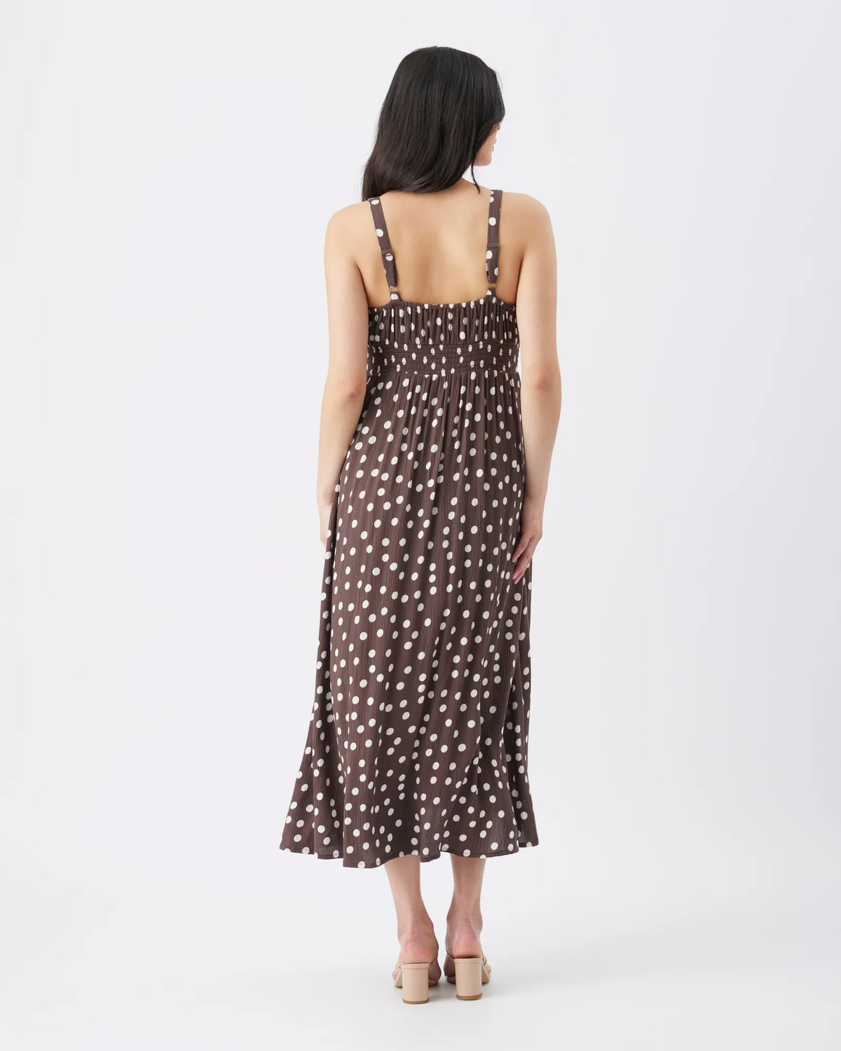 A woman viewed from the back wearing a sleeveless midi dress with thin straps and an allover white polka dot pattern on a dark brown background, paired with beige block-heel sandals, against a plain white studio background.