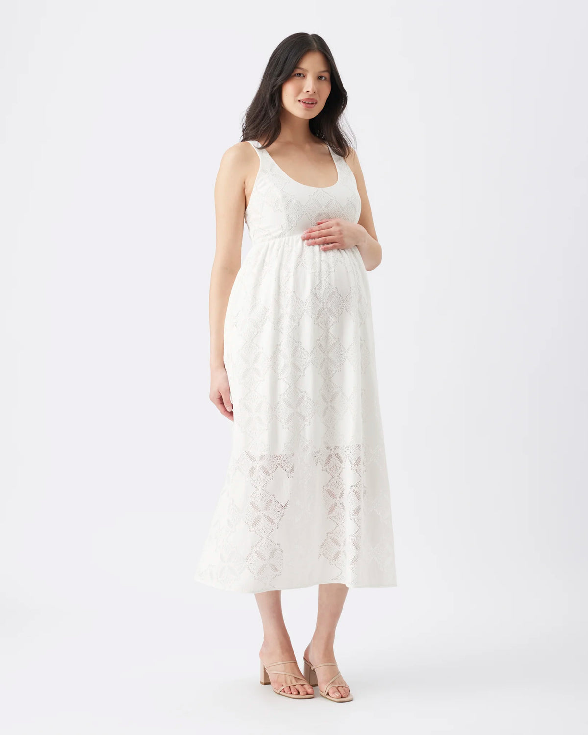 A pregnant woman modeling a sleeveless white midi dress with intricate floral lace embroidery, standing against a seamless white studio background.