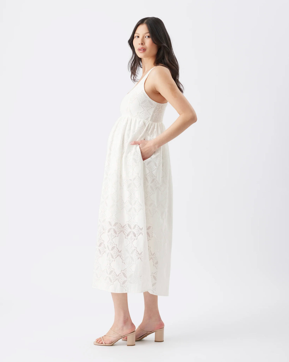 A woman modeling a sleeveless white midi dress with side pockets and delicate lace detailing near the hem, paired with beige block-heel mule sandals against a plain white studio background.