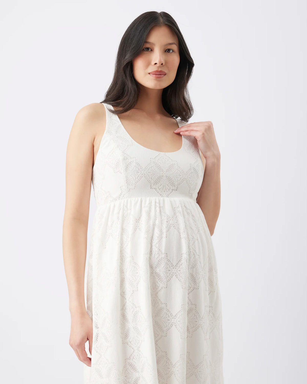 A pregnant model wearing a white sleeveless maternity dress with a textured, embroidered geometric pattern, photographed against a clean white studio background.