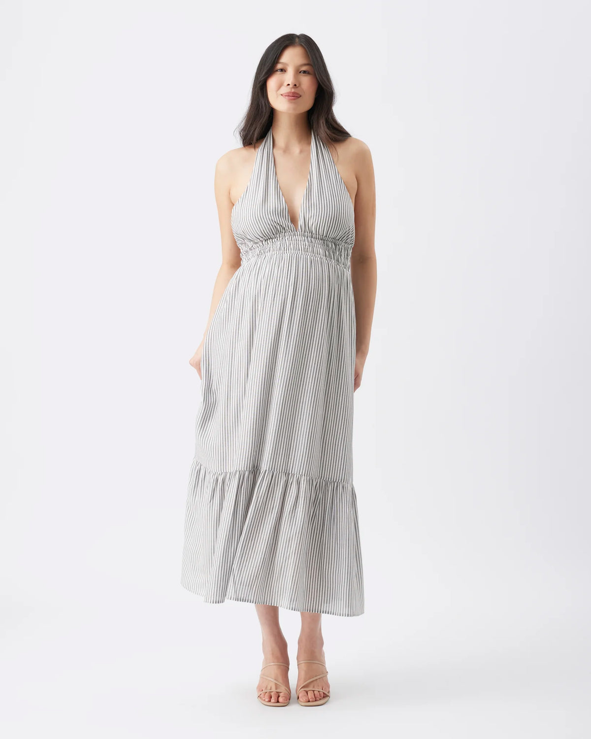 A woman modeling a sleeveless halter-neck maxi dress with vertical pinstripes in light gray and white, set against a plain white studio background.