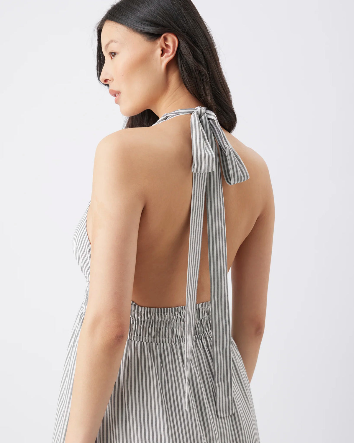 A woman shown from the back wearing a sleeveless halter dress with a tie-back neckline and a gray-and-white vertical stripe pattern, photographed against a clean light gray studio background.