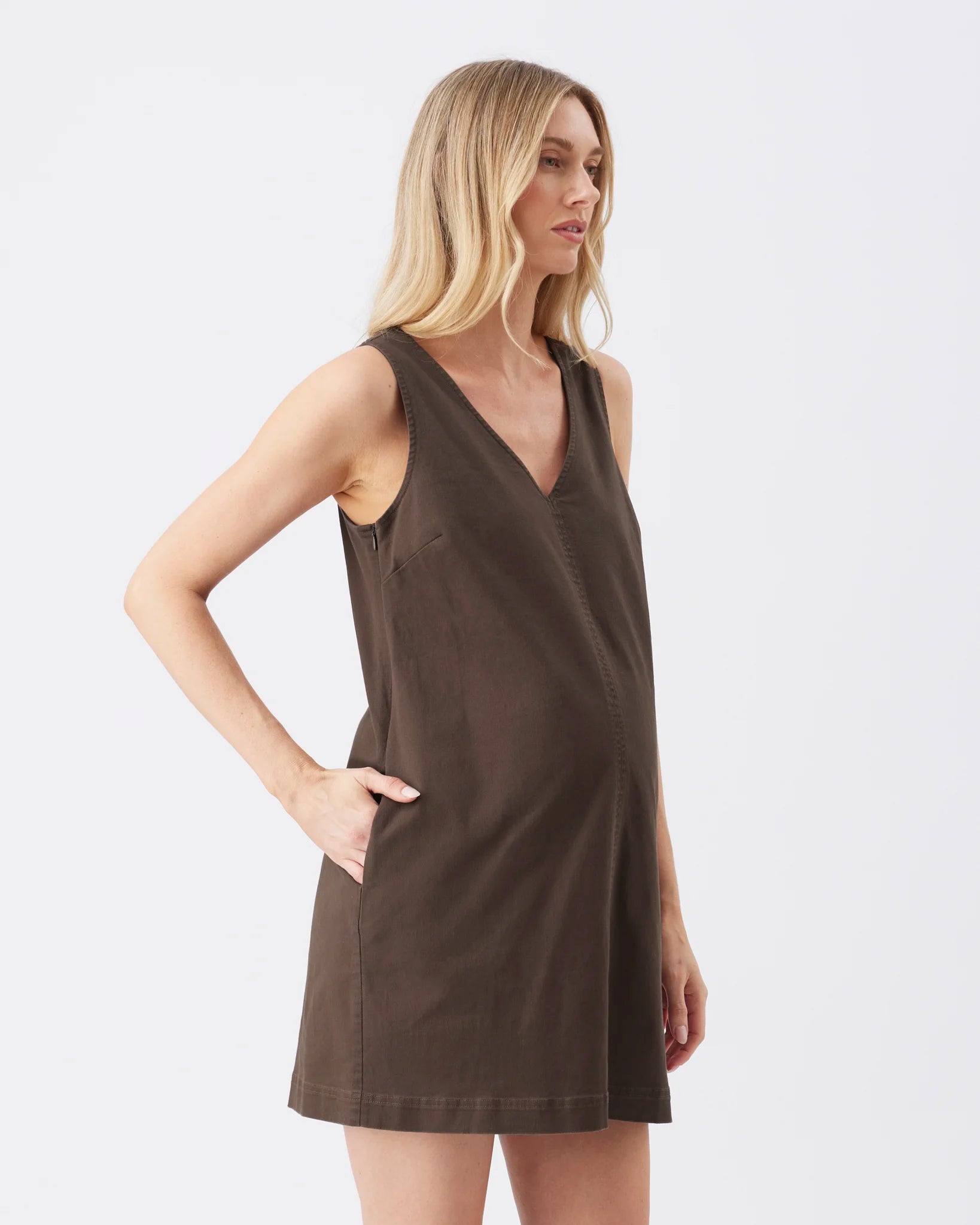 A blonde woman modeling a sleeveless, V-neck mini dress in a dark brown color, shown from a side profile against a plain white studio background.