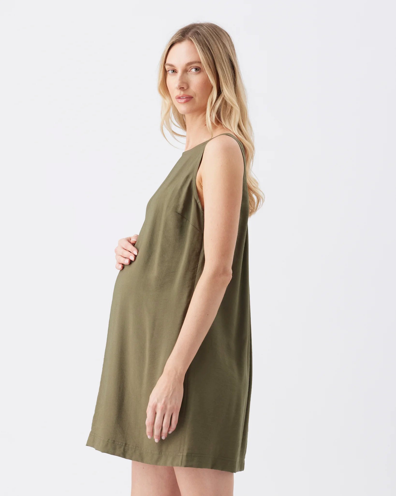 A pregnant woman wearing a sleeveless olive green maternity shift dress, shown from a side profile against a plain white studio background.