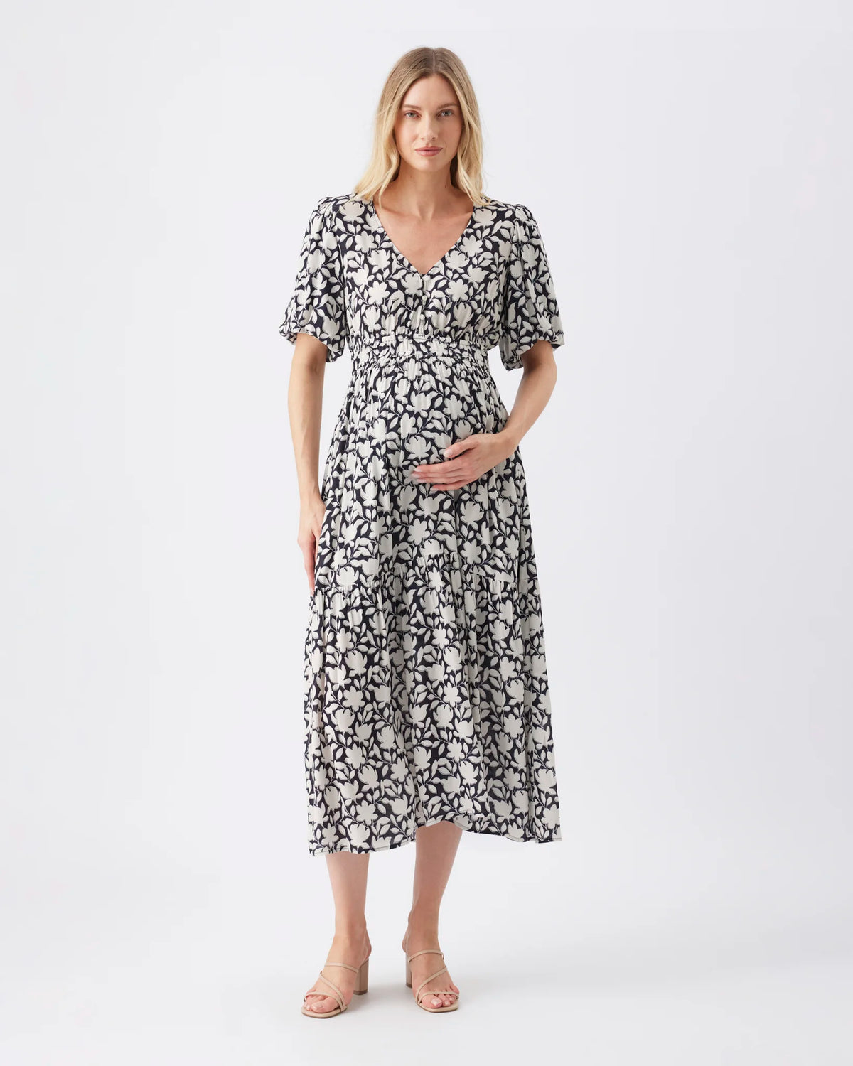 A pregnant model wearing a black and white floral print midi dress with short puff sleeves and a V-neckline, standing against a plain white studio background.