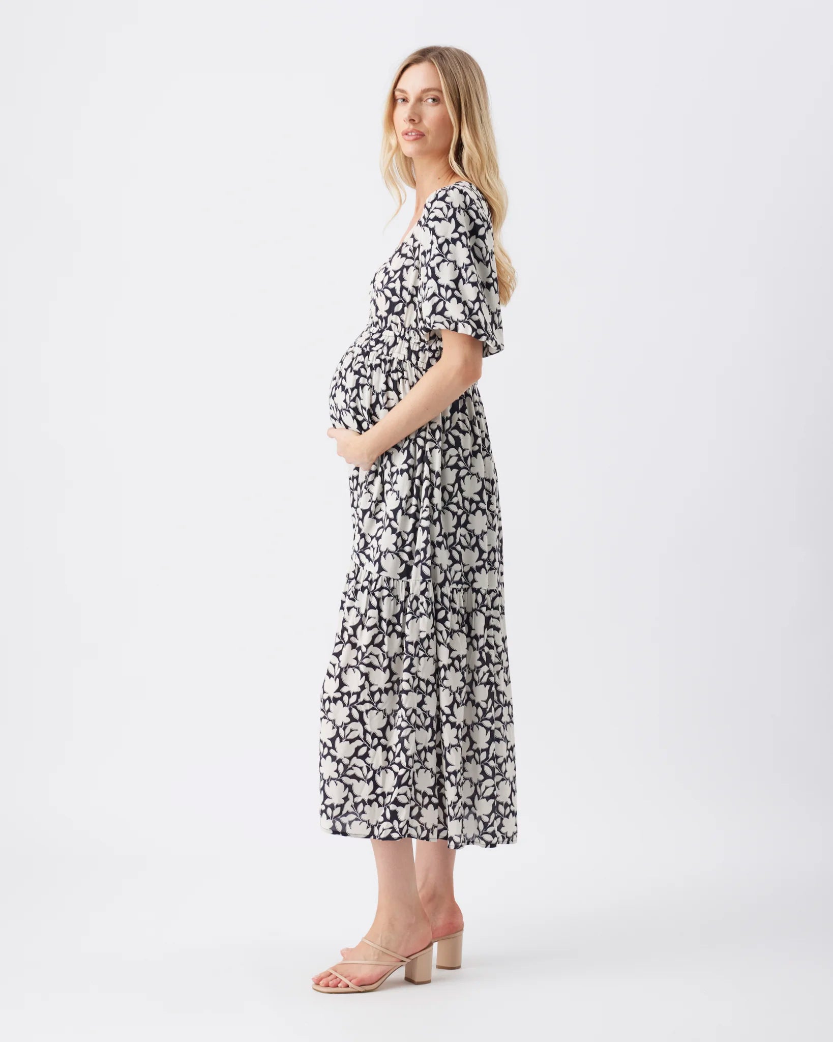 A pregnant woman wearing a black and white floral print midi dress with flutter sleeves, standing in profile against a plain white studio background.