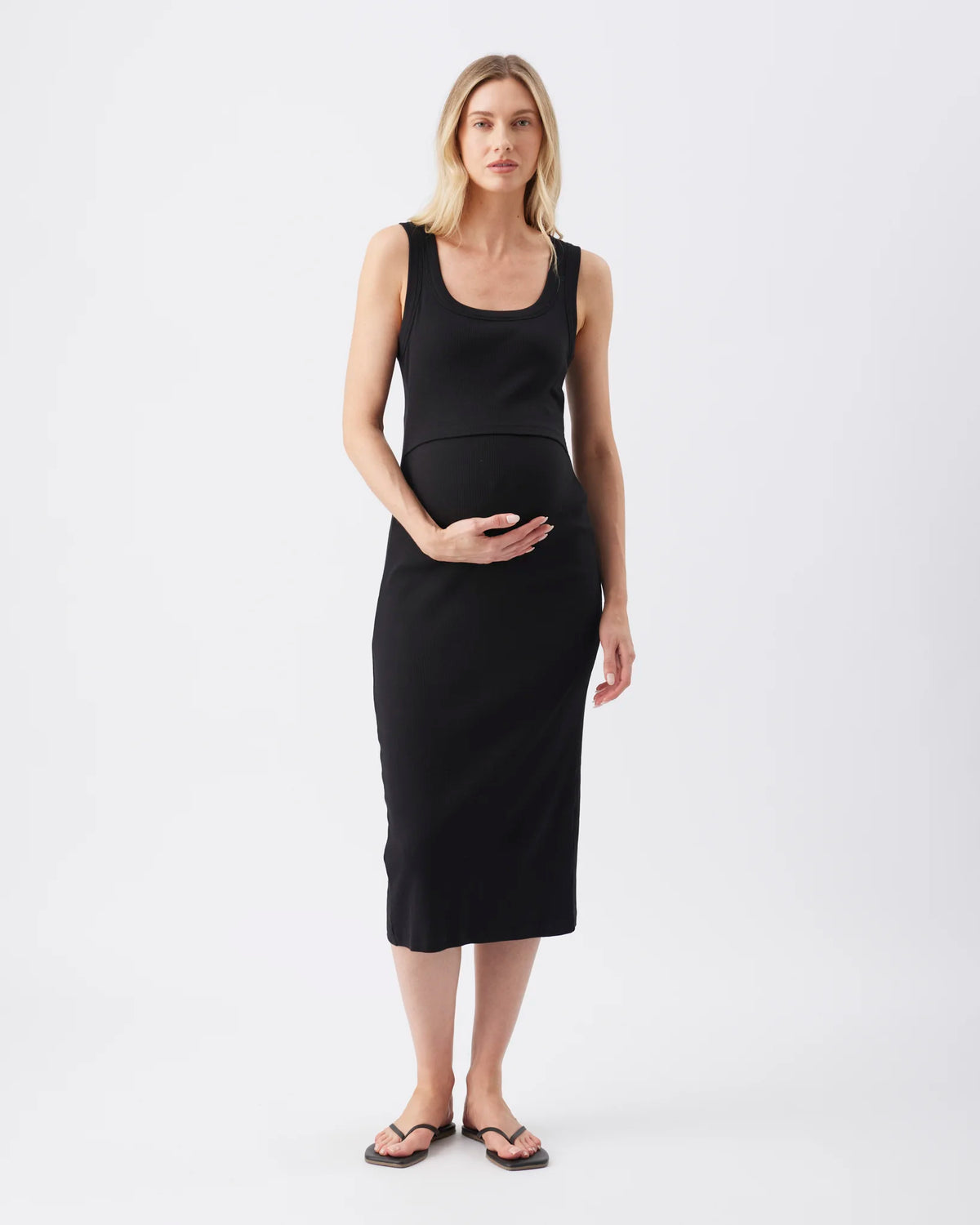 A pregnant woman modeling a black sleeveless maternity dress with a scoop neckline and a fitted silhouette, standing against a plain white studio background.