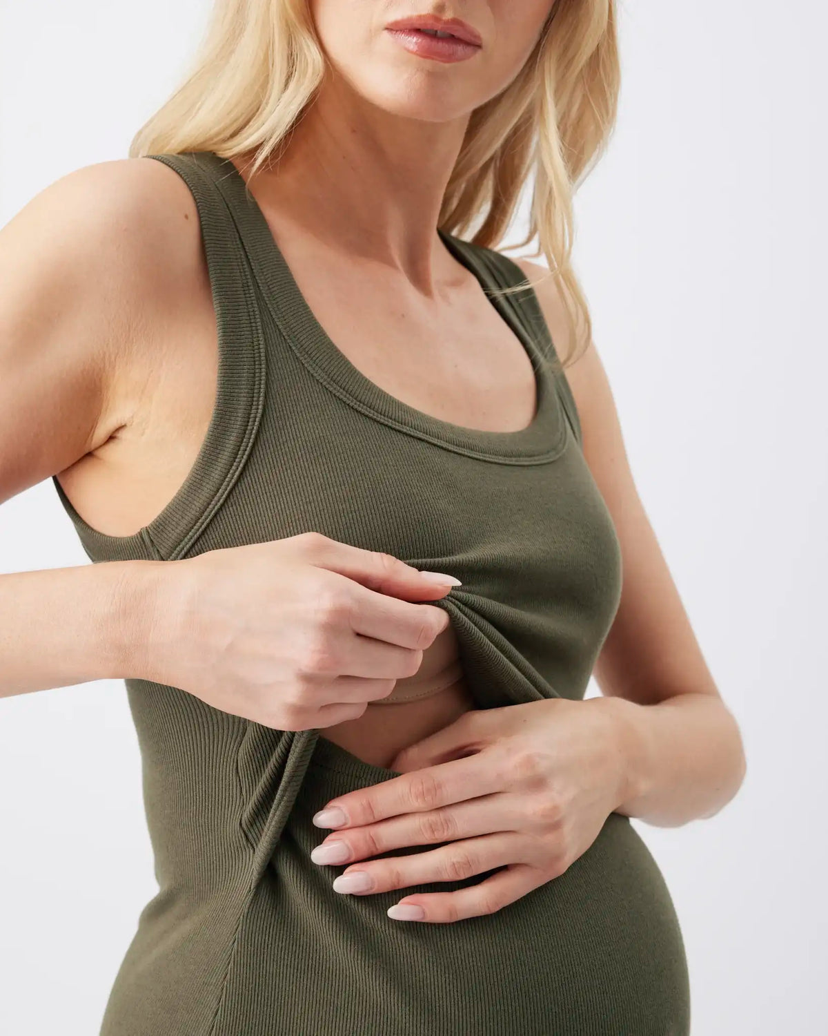 A close-up of a woman wearing an olive green ribbed tank top, lifting the fabric of the front to show its stretch and fit against a plain light background.