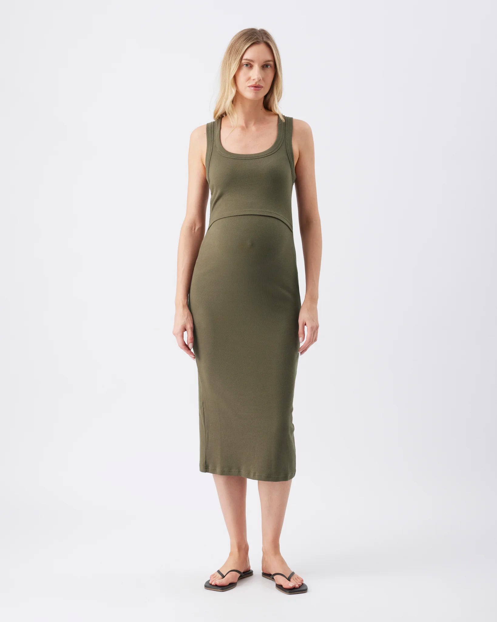 A woman modeling a sleeveless olive green maternity dress with a scoop neckline and a fitted, knee-length silhouette, standing against a plain light gray studio background.