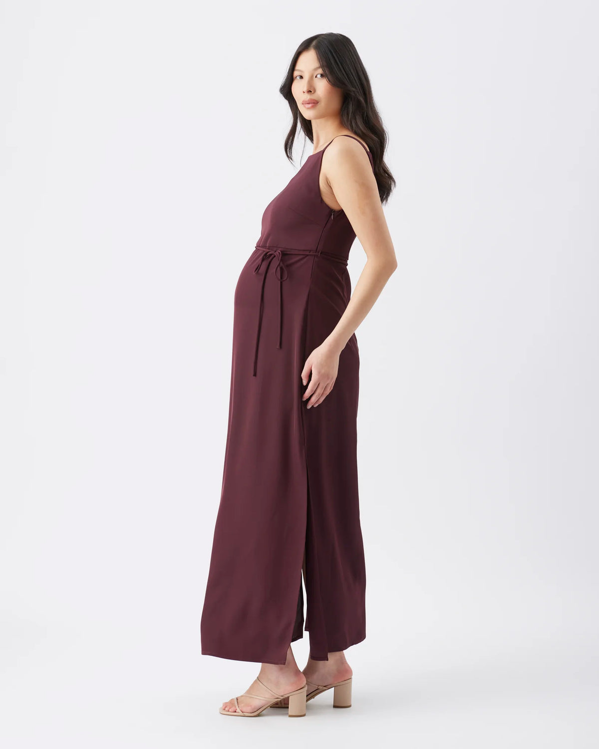 A pregnant model wearing a sleeveless, wide-leg burgundy maxi jumpsuit with a tie waist, standing in a side profile against a light gray studio background.