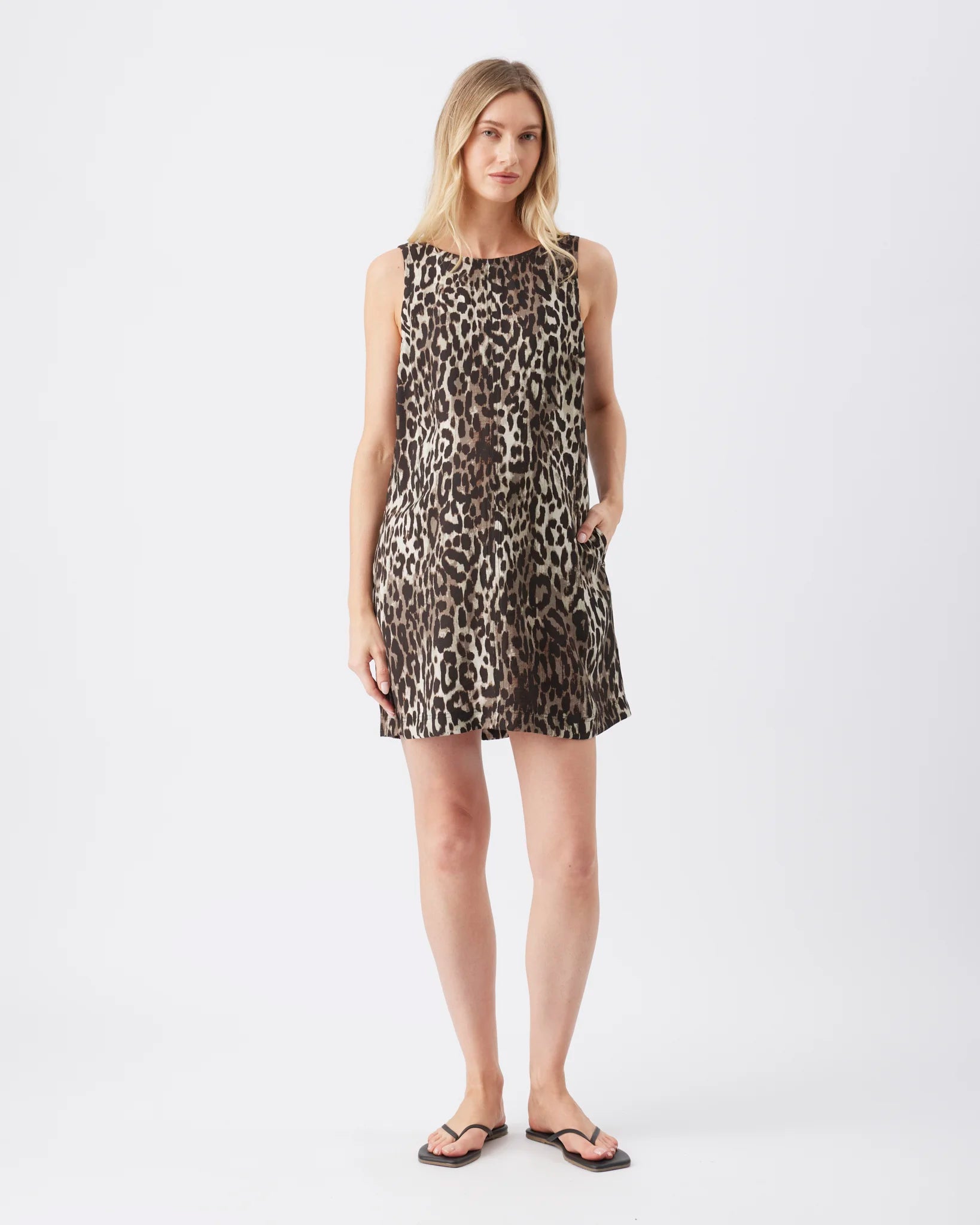 A woman modeling a sleeveless leopard-print mini dress with a relaxed fit, paired with dark flip-flop sandals, against a plain light gray studio background.