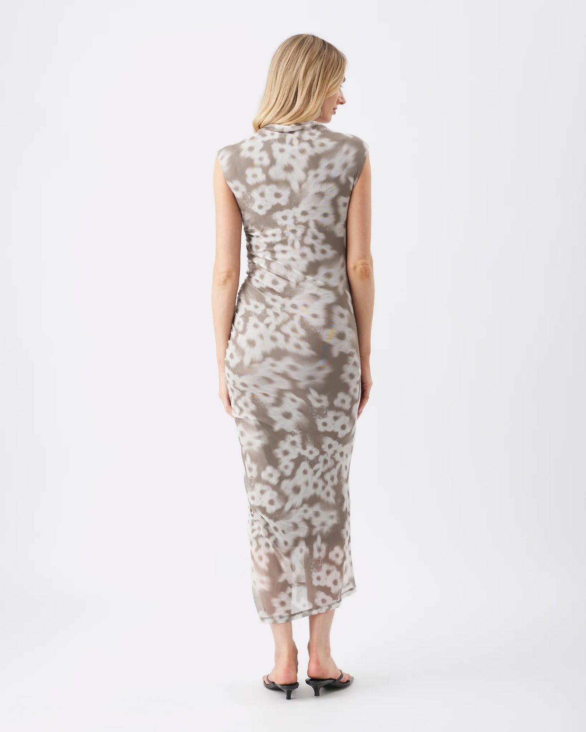A woman viewed from the back wearing a sleeveless, knee-length fitted dress with an abstract floral or organic print in beige and gray tones, paired with black flat sandals, against a plain white studio background.