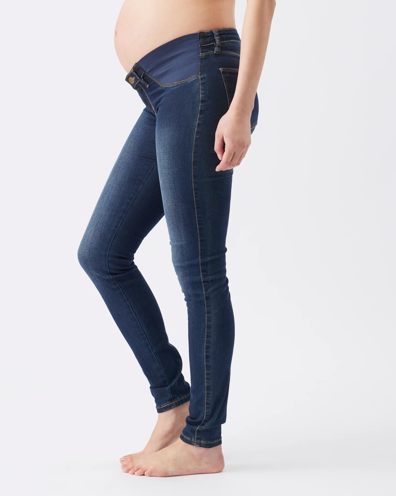 A side-profile view of a pregnant model wearing dark blue maternity skinny jeans against a plain white studio background.