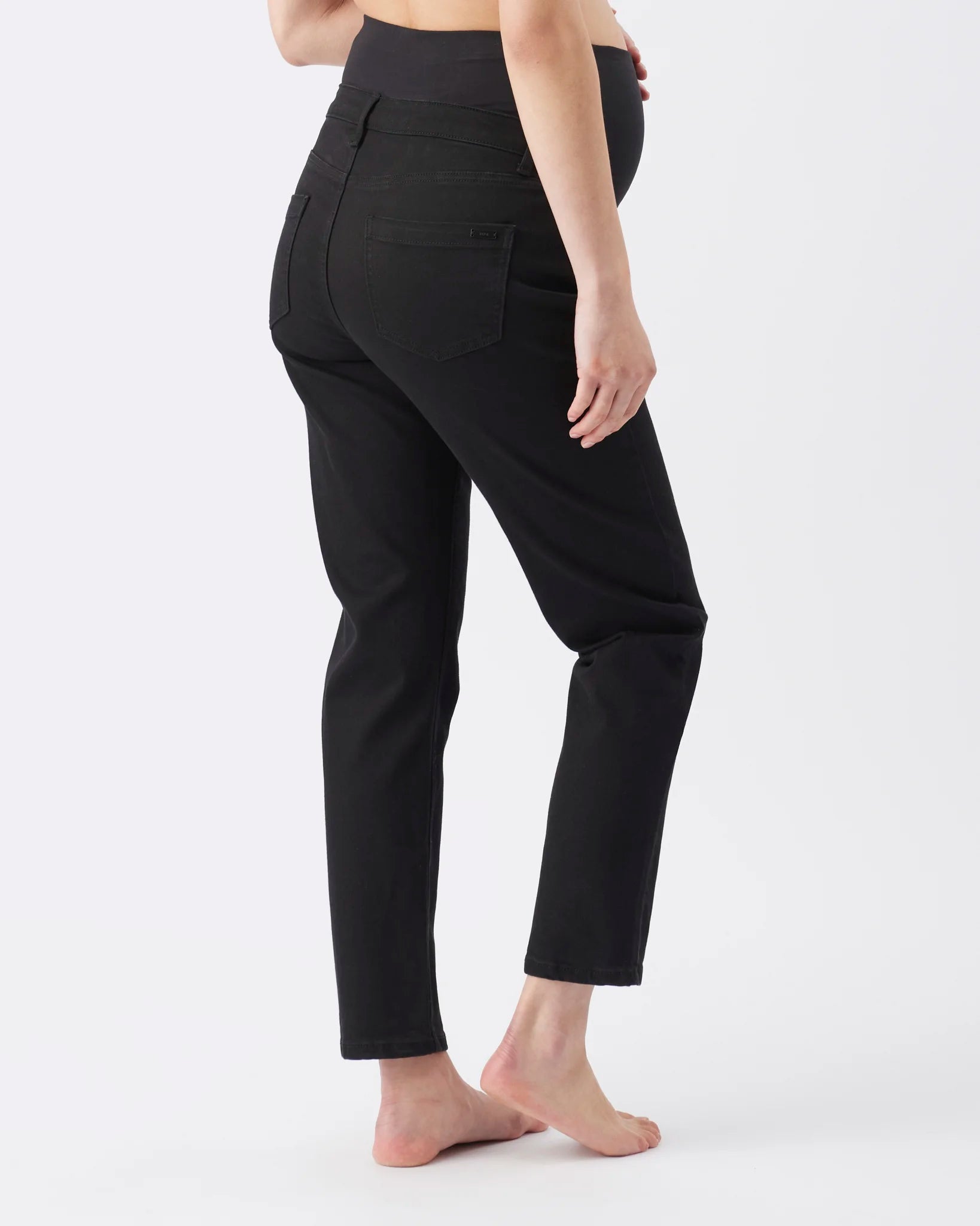 A person shown from the back wearing black high-waisted cropped pants, standing barefoot against a plain light gray studio background.