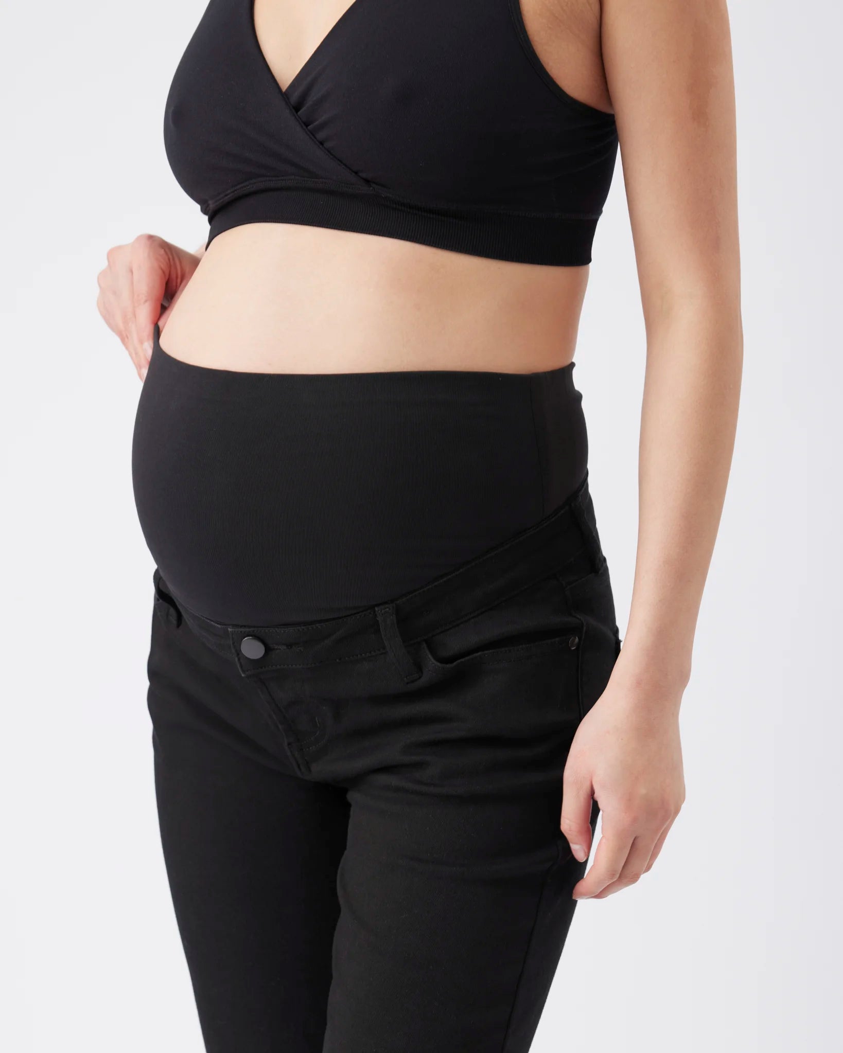 A close-up of a pregnant model wearing a black maternity support bra and black high-waisted jeans with a built-in maternity belly band.