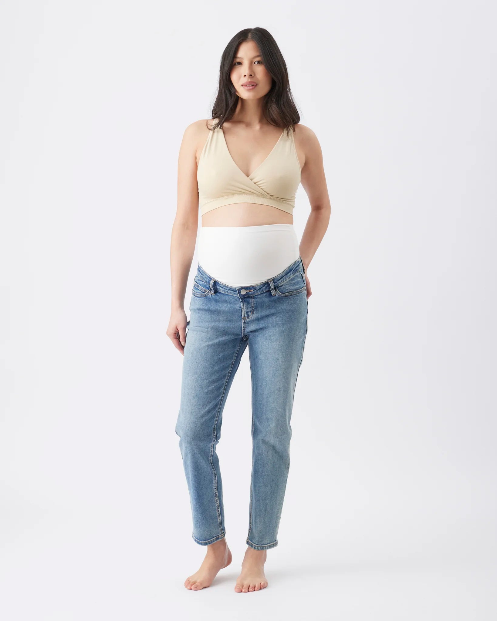 A pregnant woman stands barefoot against a light gray studio background, wearing a beige wrap-style maternity top and light blue jeans.