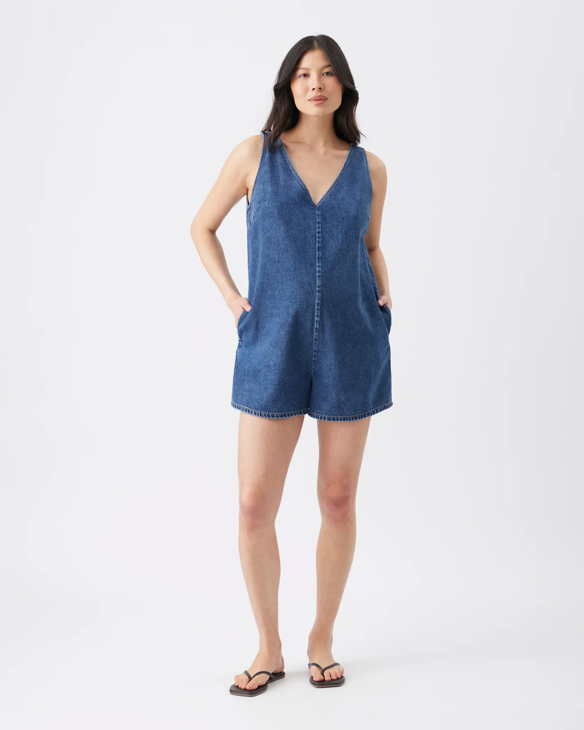A woman modeling a sleeveless blue denim romper with a V-neckline, front button detail, and side pockets, paired with dark flip-flop sandals against a plain light gray studio background.