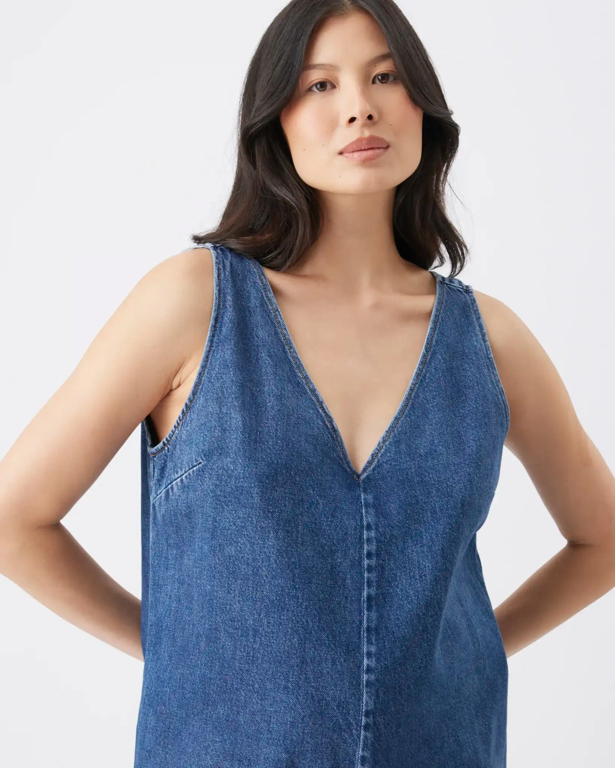 A woman wearing a sleeveless blue denim top with a deep V-neck and button-front detail, photographed against a clean light gray studio background.
