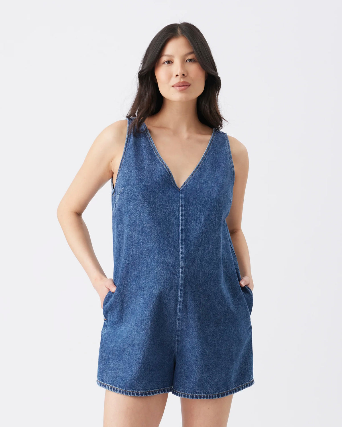 A woman modeling a sleeveless blue denim romper with a V-neckline and front pockets, photographed against a plain light gray studio background.