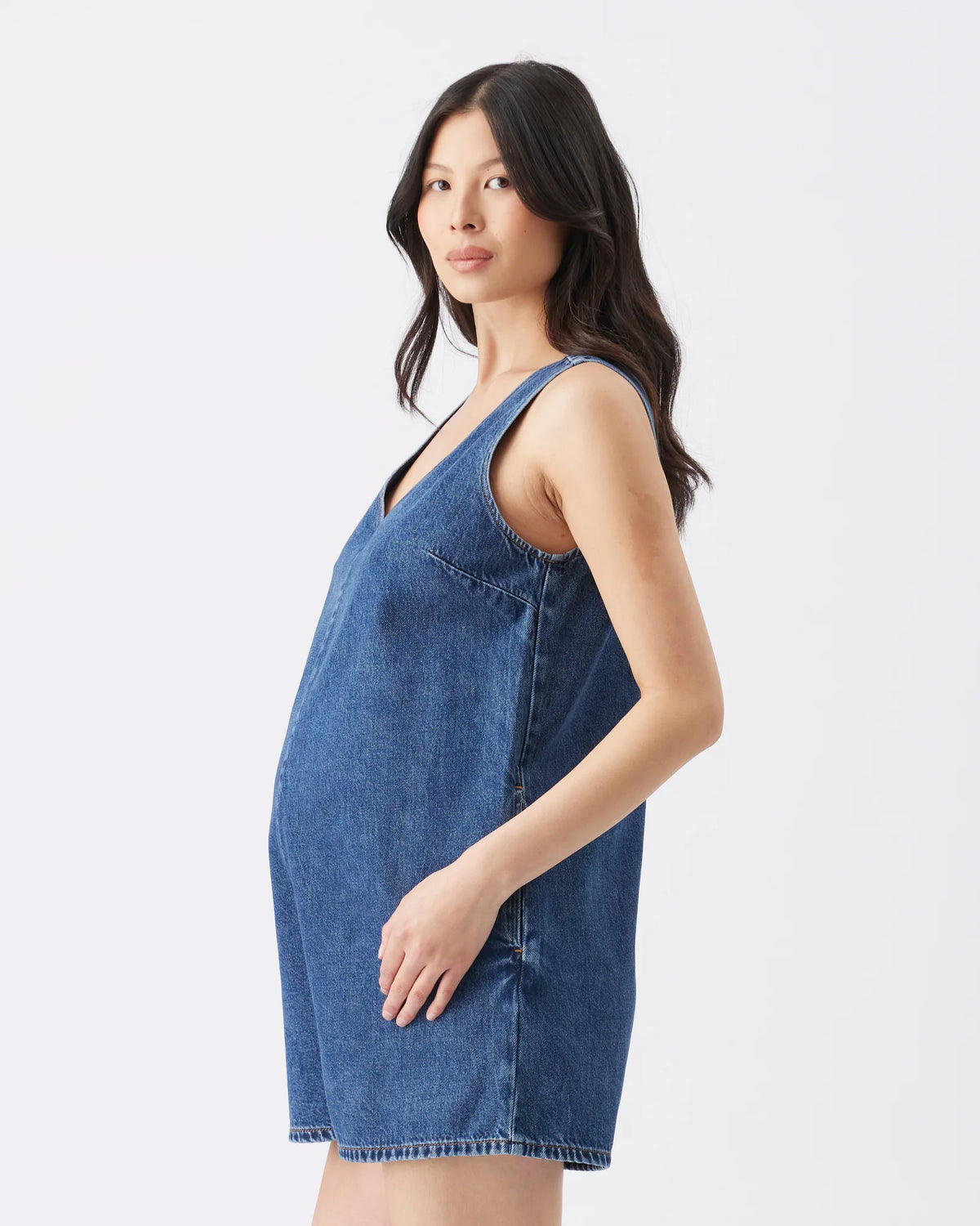 A pregnant model wearing a blue denim maternity dress, shown from a side profile against a plain white studio background.