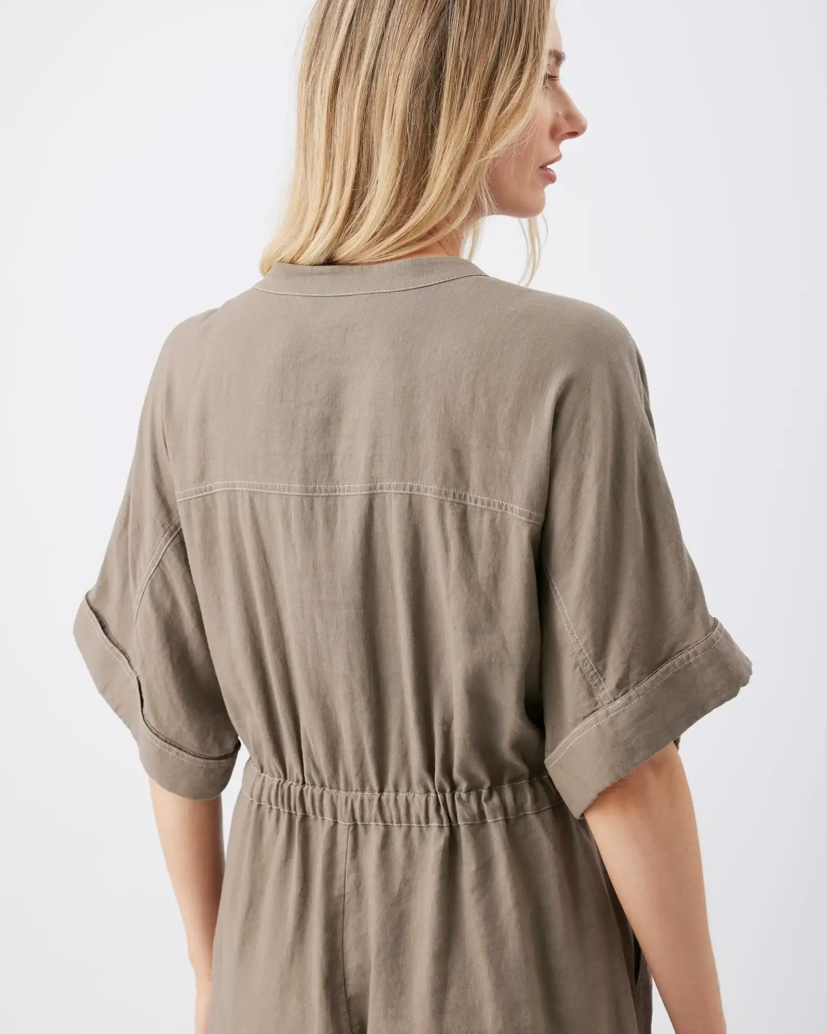 A woman shown from the back wearing a loose, short-sleeve taupe dress with rolled cuffs and a gathered elastic waist, photographed against a plain light background.