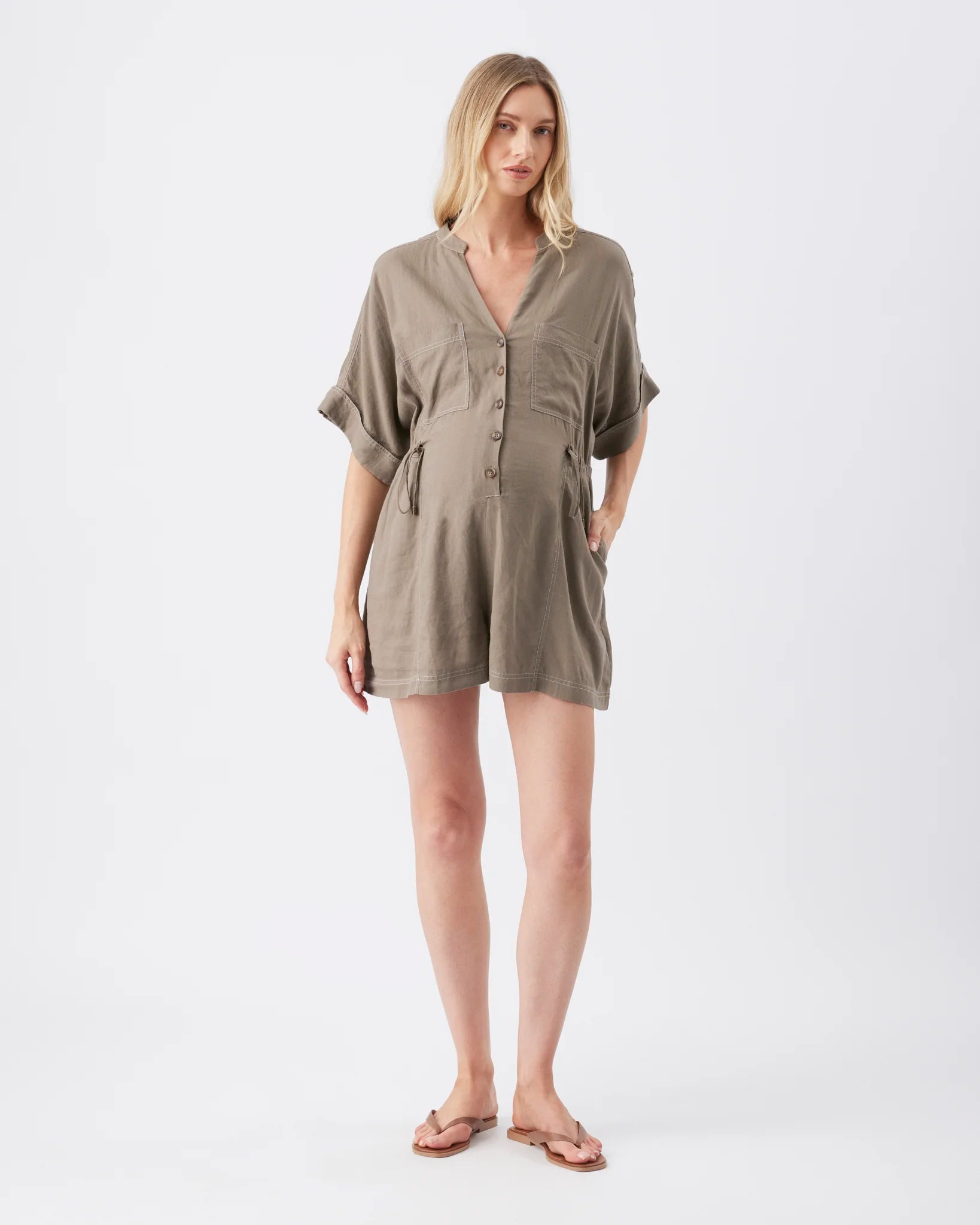 A pregnant model wearing a short-sleeved, button-front linen romper in a muted taupe-brown color, styled with brown sandals against a plain white studio background.