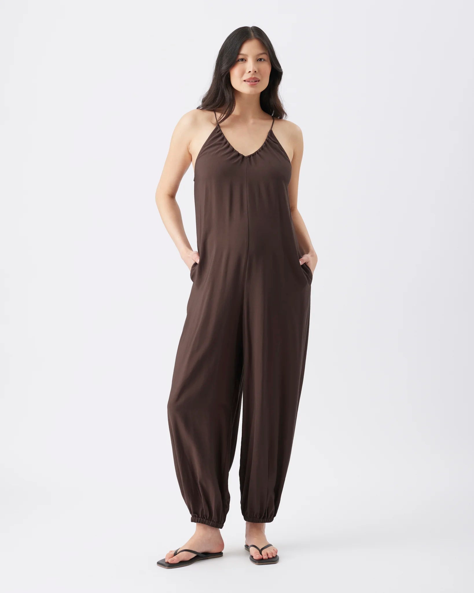 A woman modeling a dark brown sleeveless jumpsuit with a gathered halter-style neckline and wide, baggy harem-style legs with ankle cuffs, standing against a light gray studio background.