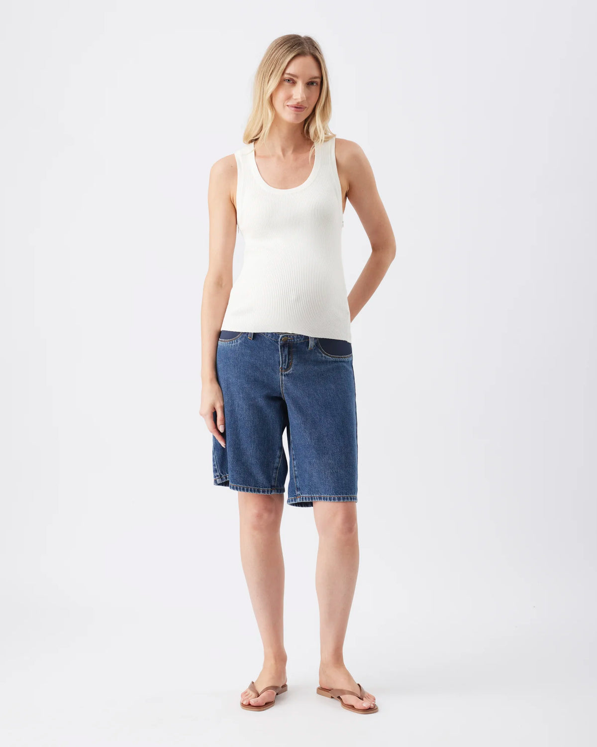 A woman modeling a white ribbed tank top and blue denim shorts against a light gray studio background.