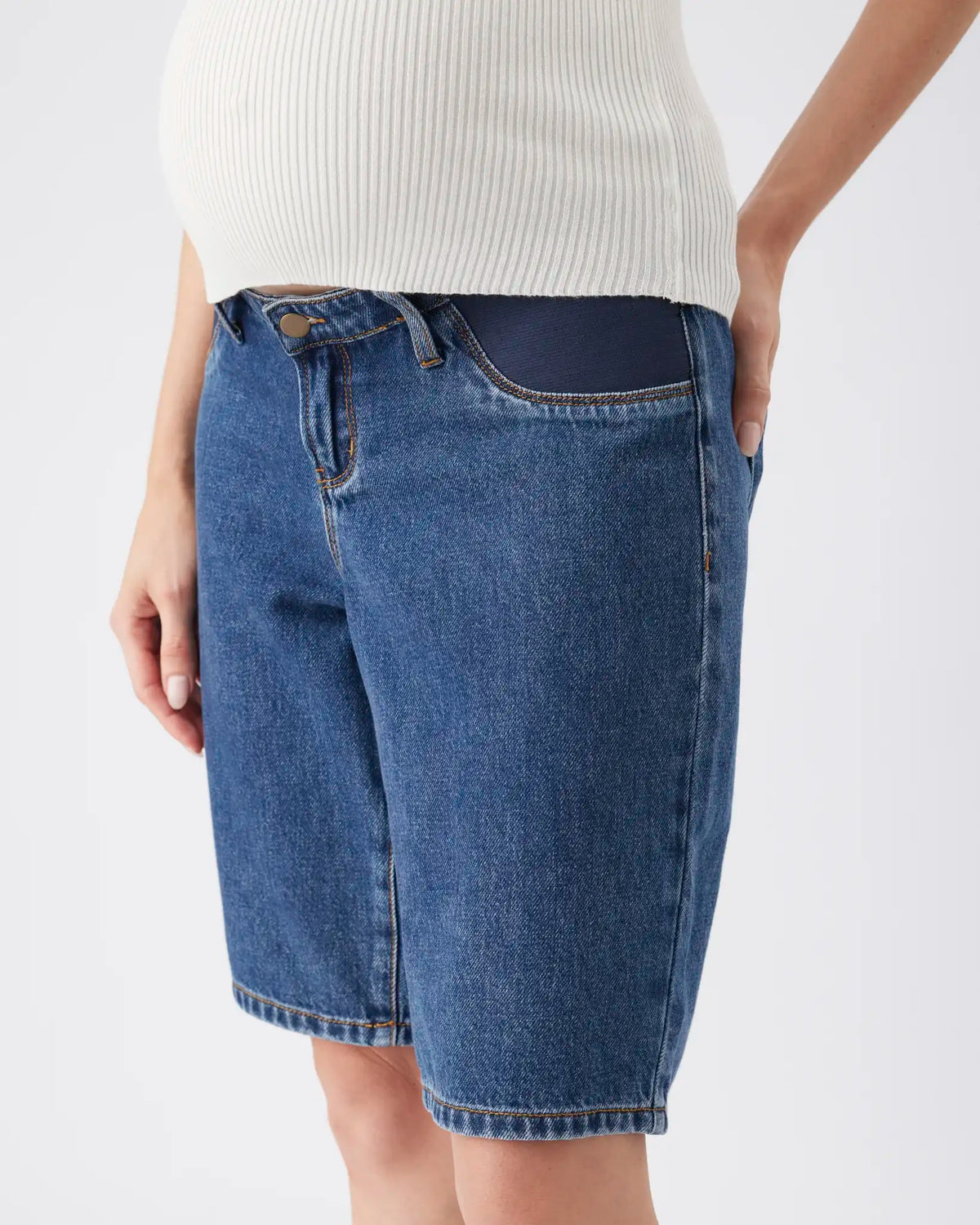 A close-up of a model wearing blue denim shorts with a dark navy contrast waistband, paired with a white ribbed knit top, against a plain light gray background.