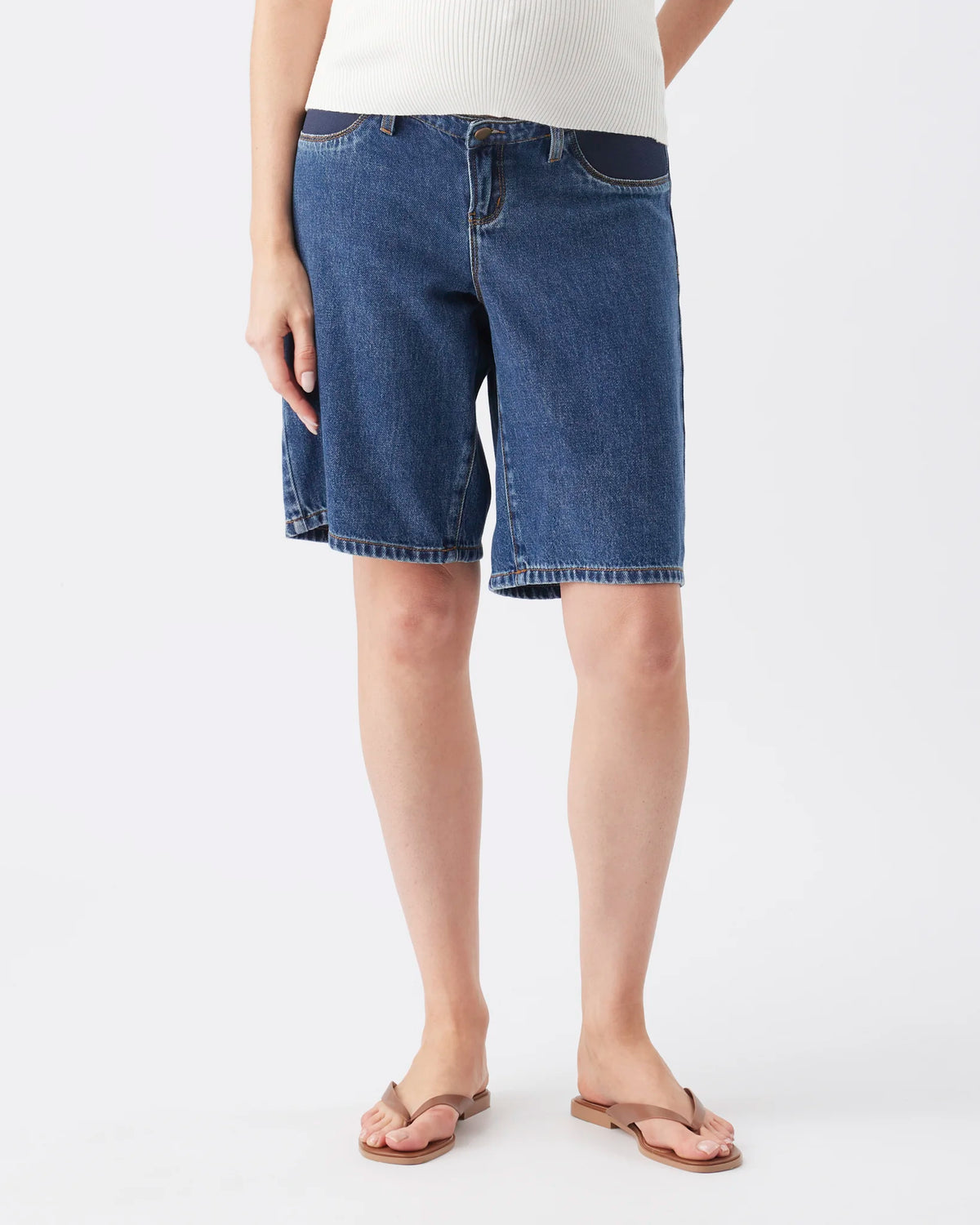A person wearing medium-wash blue denim shorts with a white ribbed top and brown flip-flop sandals, shown from the waist down against a light gray studio background.
