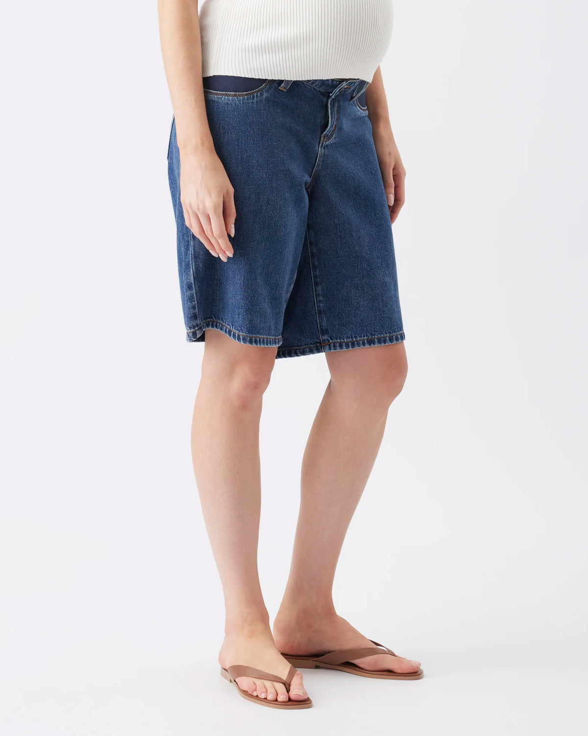 A model wearing dark blue denim shorts, a white ribbed top, and brown flip-flop sandals, shown from the waist down against a light gray studio background.