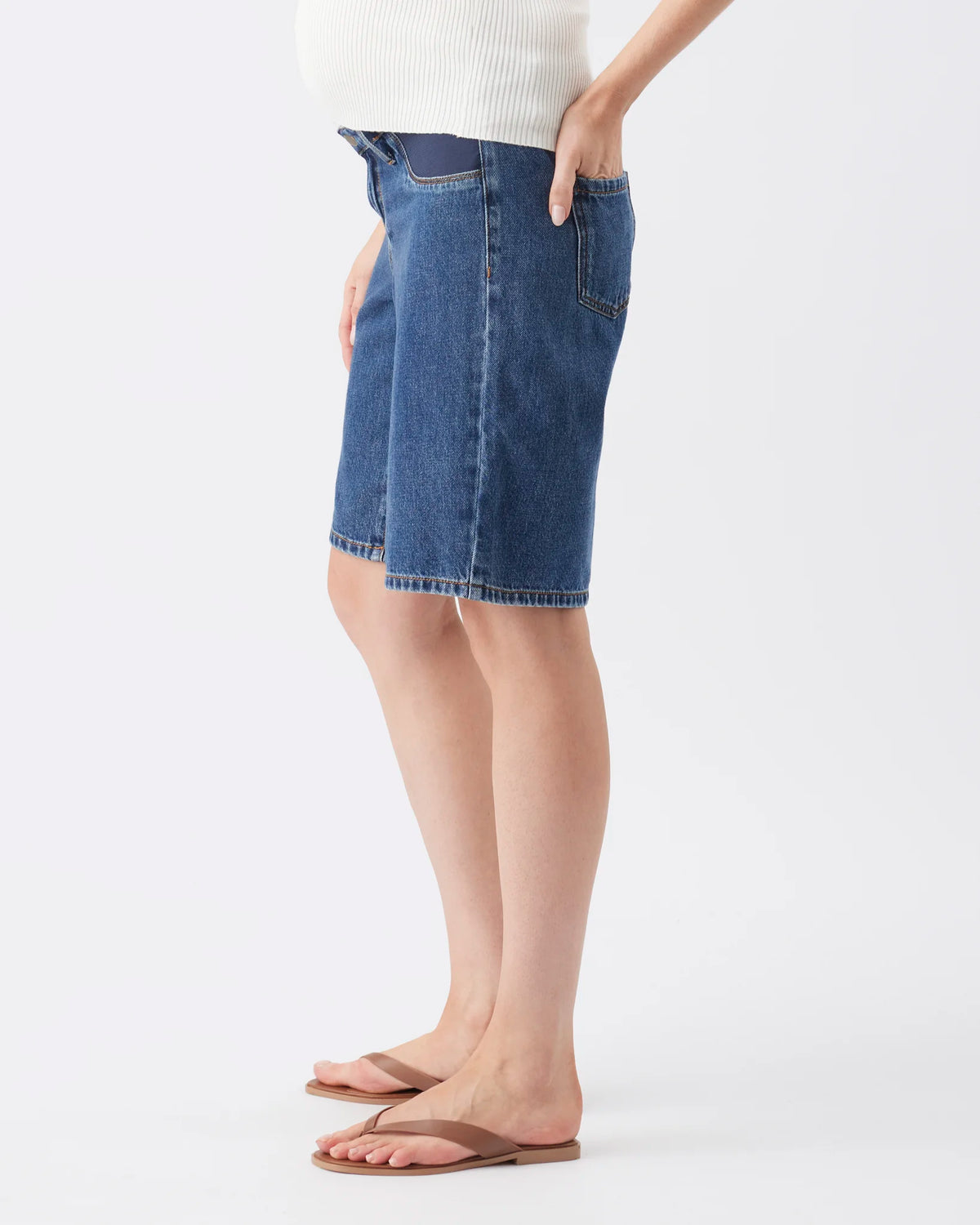 A model wearing a blue denim mini skirt with a raw hem, paired with a white ribbed top and brown flip-flop sandals, shown from the waist down against a light gray studio background.