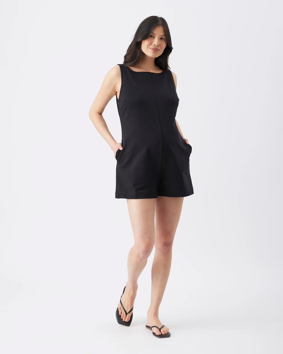A woman modeling a sleeveless black romper with side pockets, paired with black flip-flop sandals, against a plain white studio background.